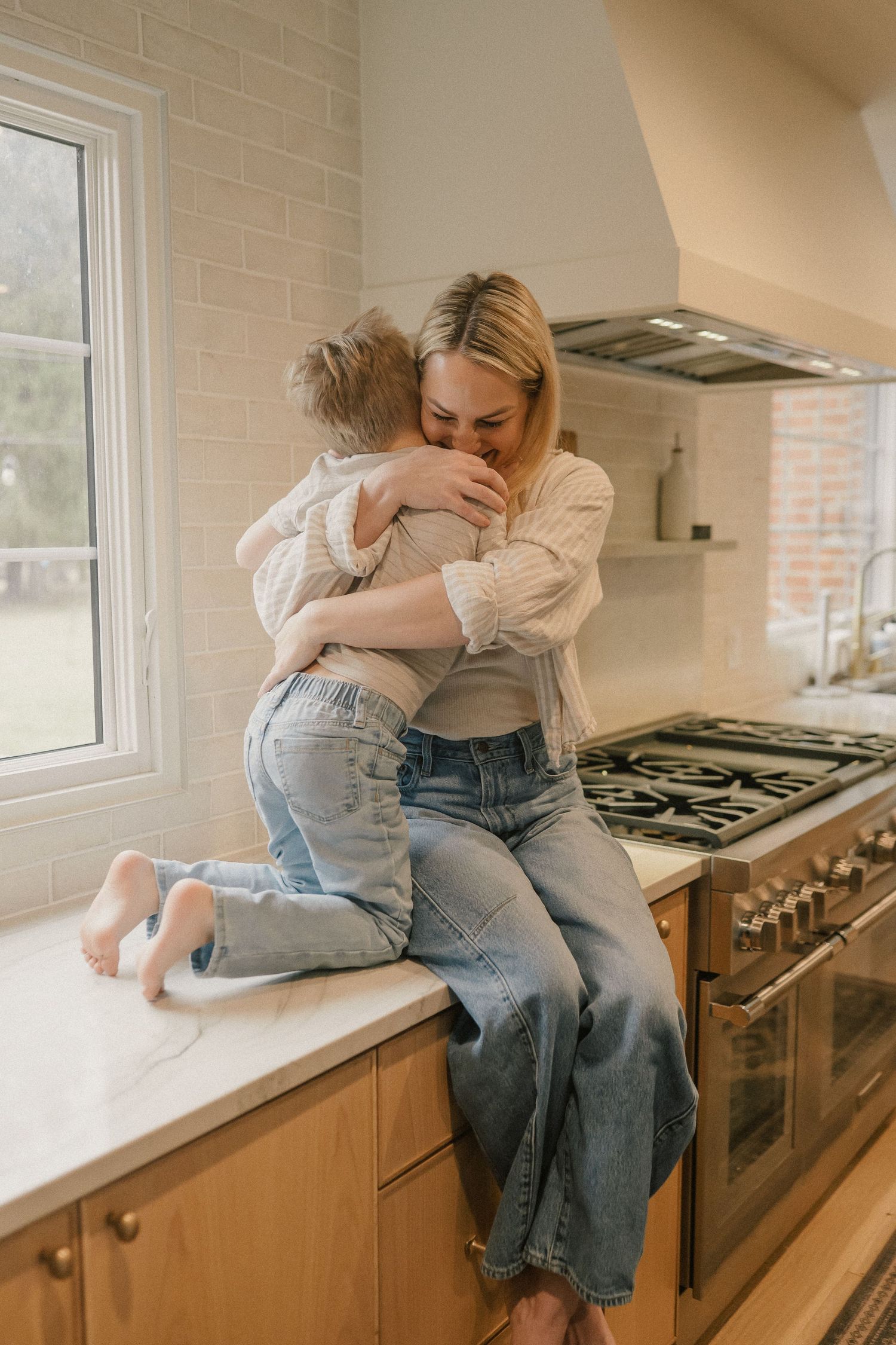 A mother and young child share a warm embrace while sitting on a kitchen counter near a gas stove.