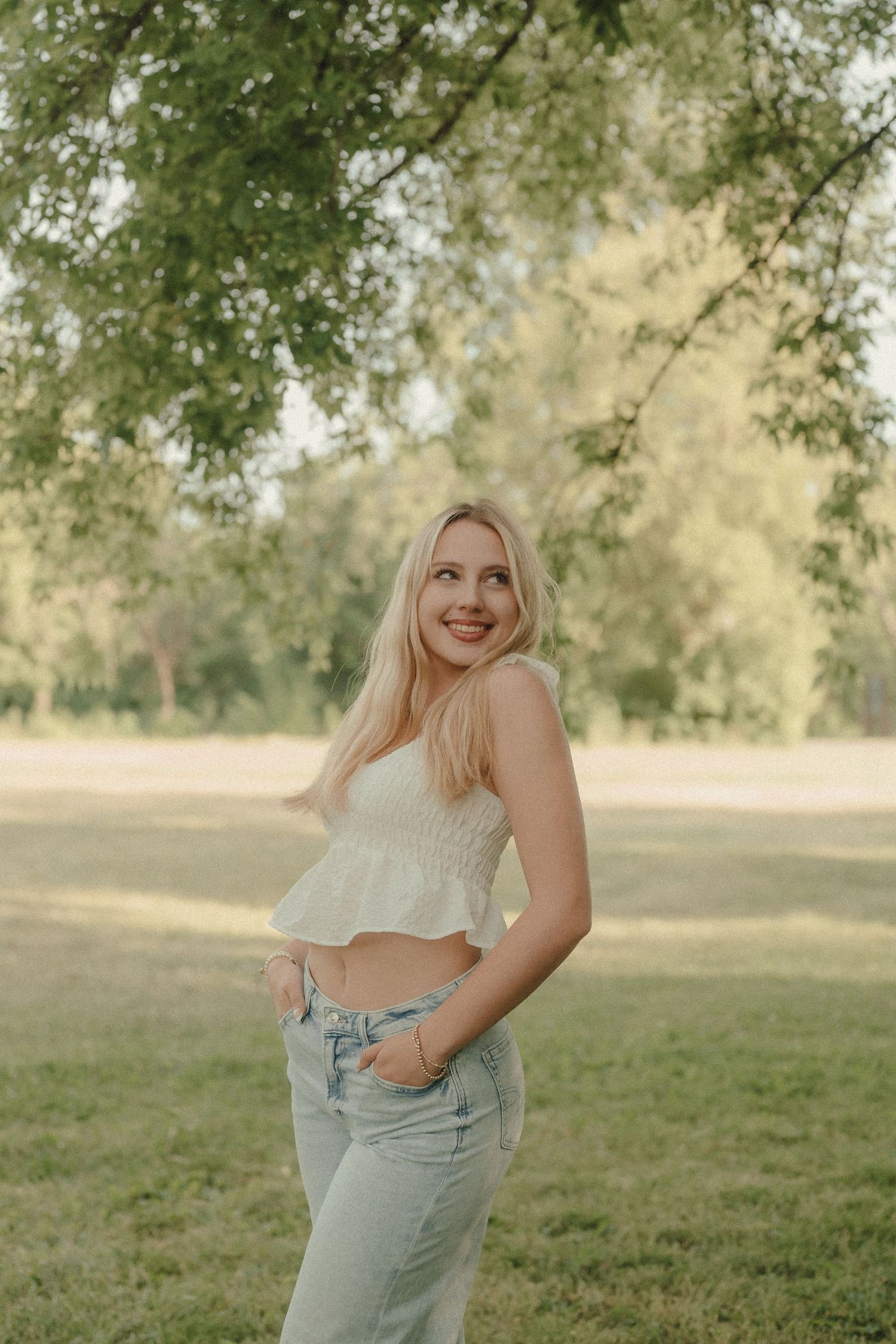 Young blonde woman in white crop top and jeans smiling outdoors in a sunny green park.