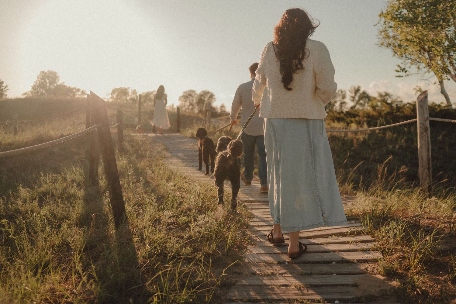 Family walking on a wooden path through a sunny meadow at golden hour, viewed from behind.