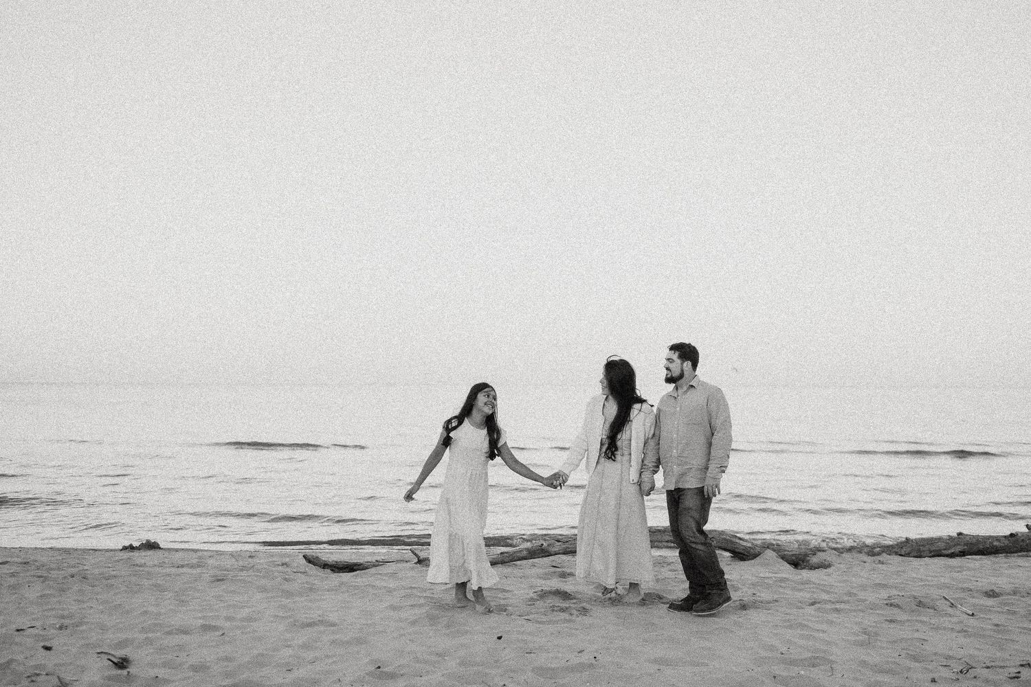 Black and white photo of three people holding hands on a sandy beach near the ocean at dusk.