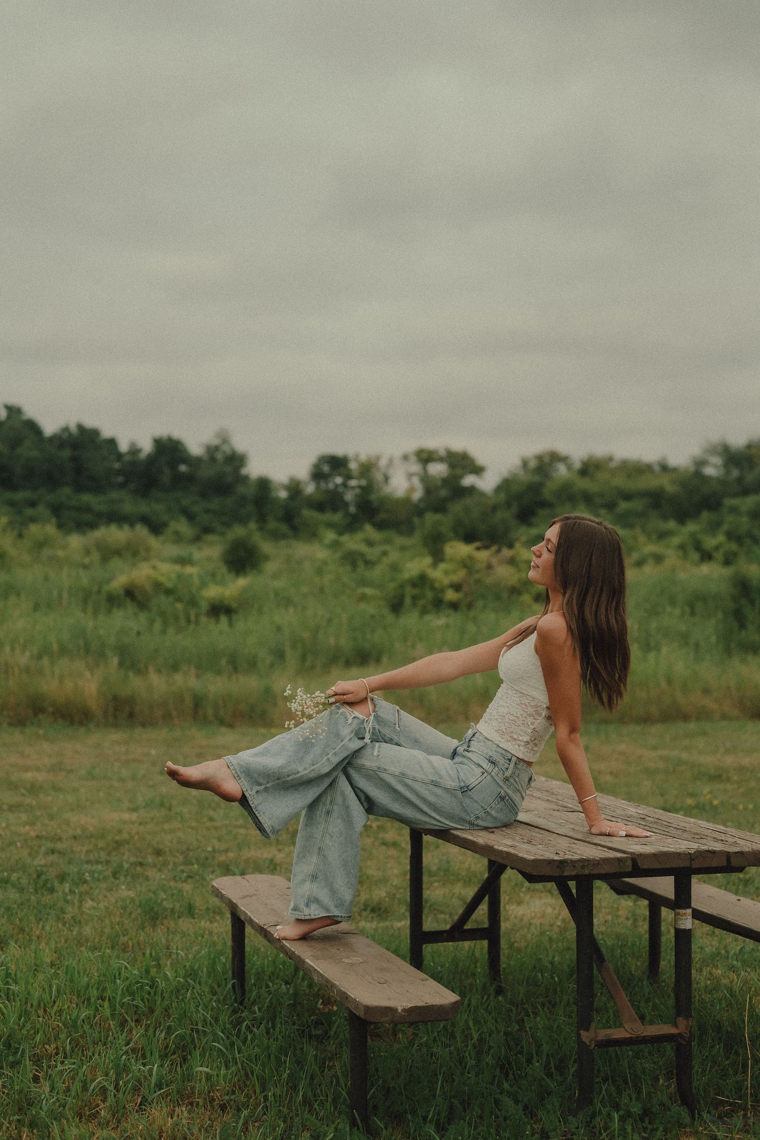 Young woman in white tank top and jeans relaxing on a picnic table in a lush green countryside.
