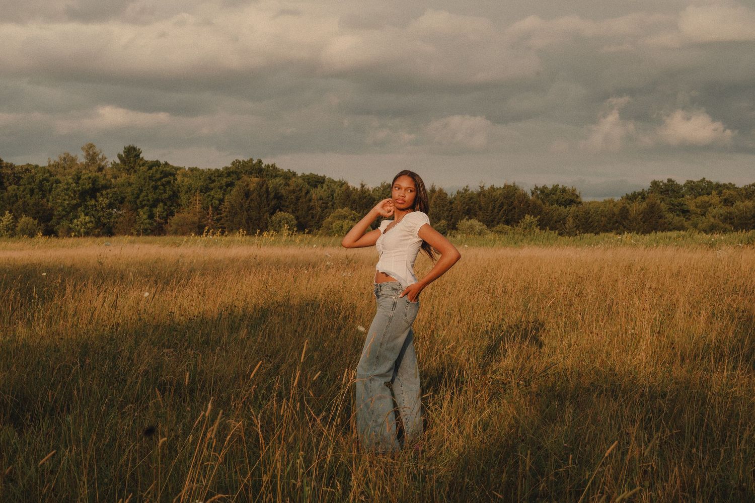 Young woman in white top and jeans posing in golden meadow with dramatic cloudy sky and forest backdrop.
