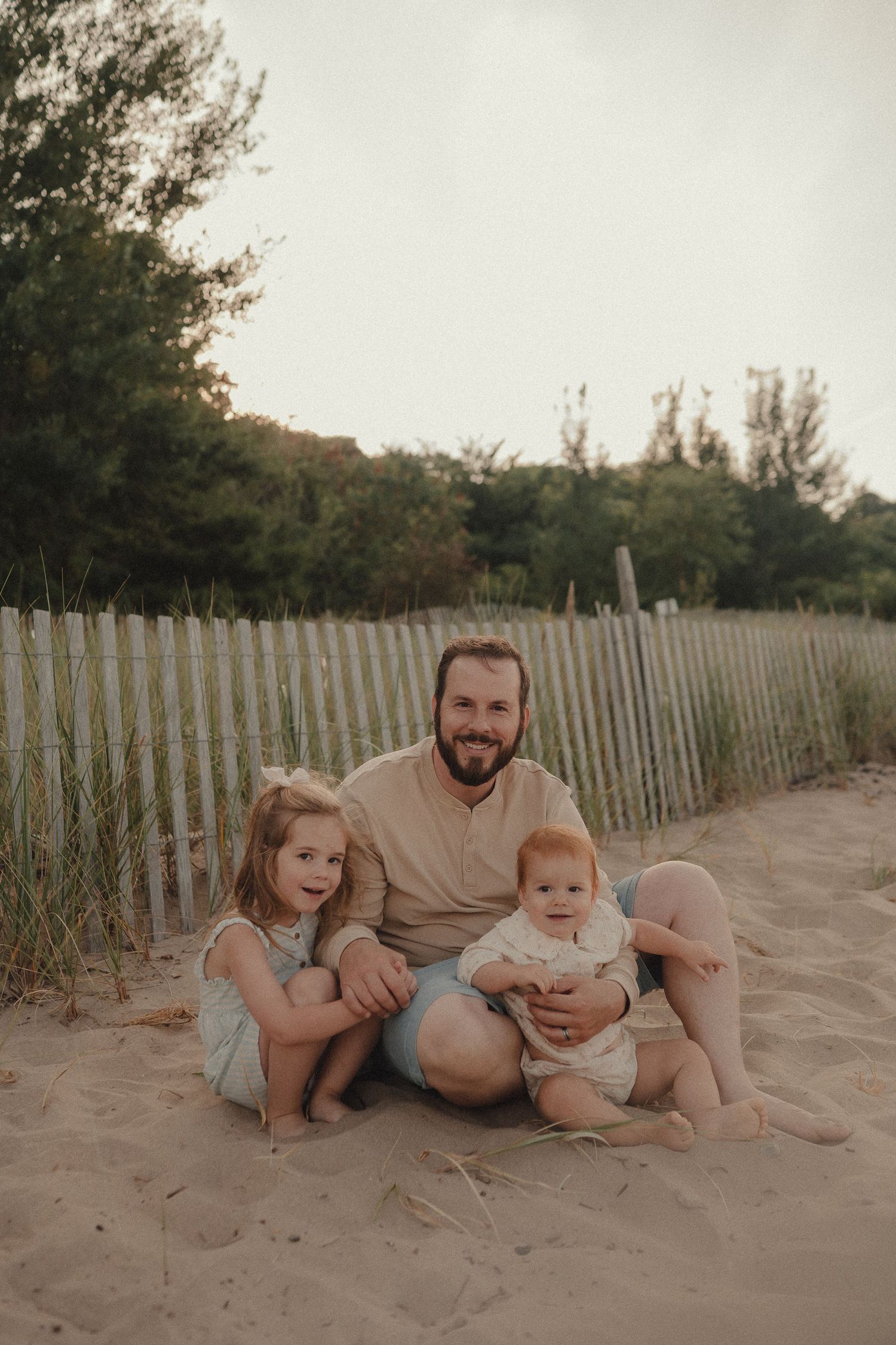 Father sitting on sandy beach with two young daughters, smiling near wooden fence with green trees.