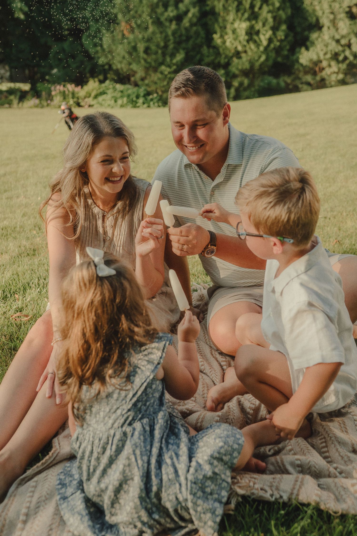 Joyful family of four sitting on blanket in sunny park sharing popsicles and laughing together.