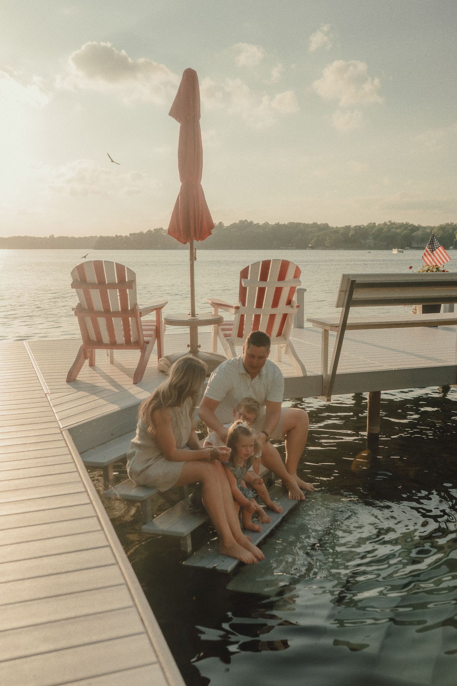A family sits together on a lakeside dock at sunset, with Adirondack chairs and an American flag in the background.