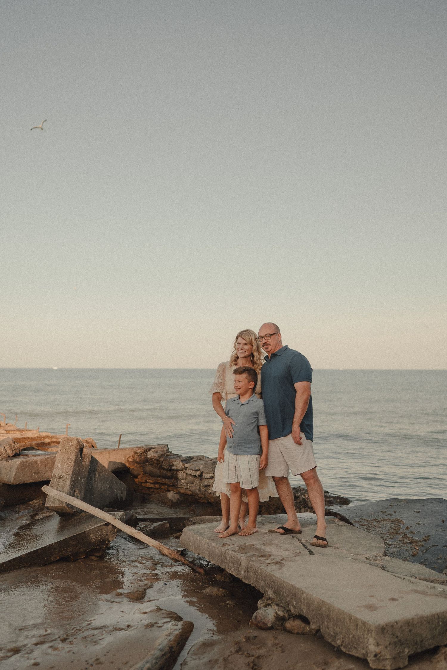 Family of four posing on rocky shoreline at sunset with calm ocean waters in the background.