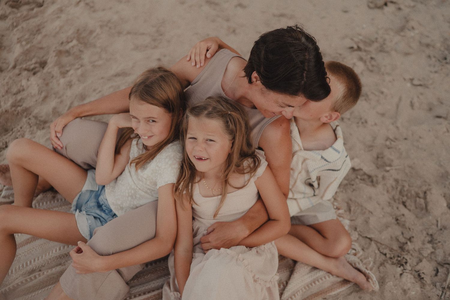 Mother cuddles three laughing children on a sandy beach blanket outdoors.