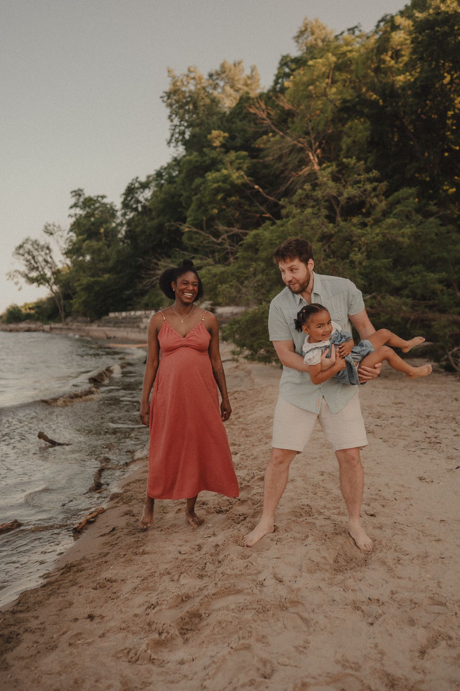Joyful interracial family on beach, father lifting laughing toddler while pregnant mother watches in coral dress.