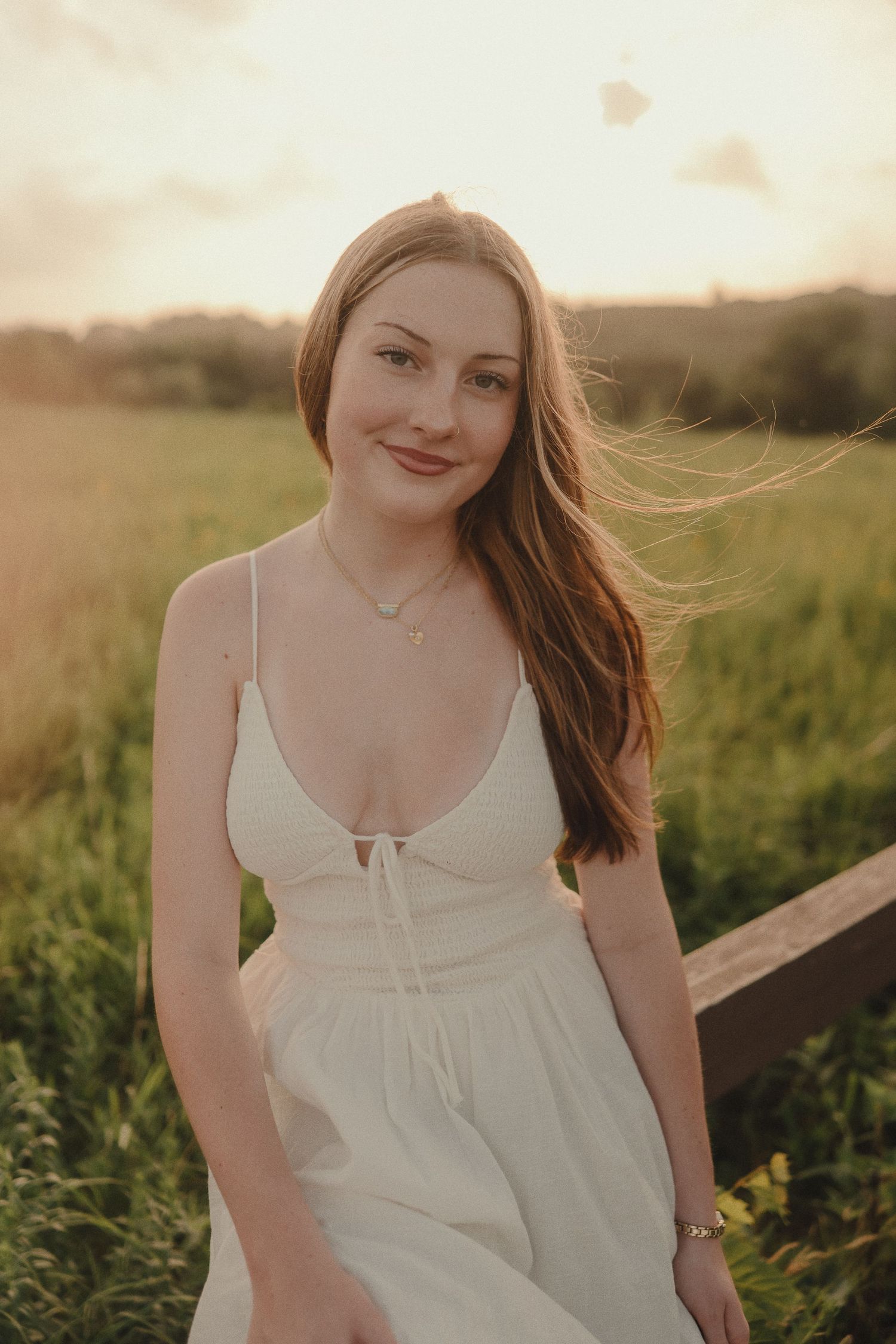 Young woman in white sundress sitting on a fence in a green field during golden hour sunset.