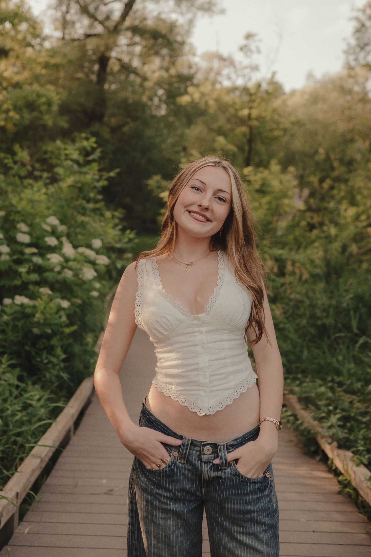 Young woman in white lace crop top and jeans smiling on a wooden boardwalk path surrounded by lush green foliage.