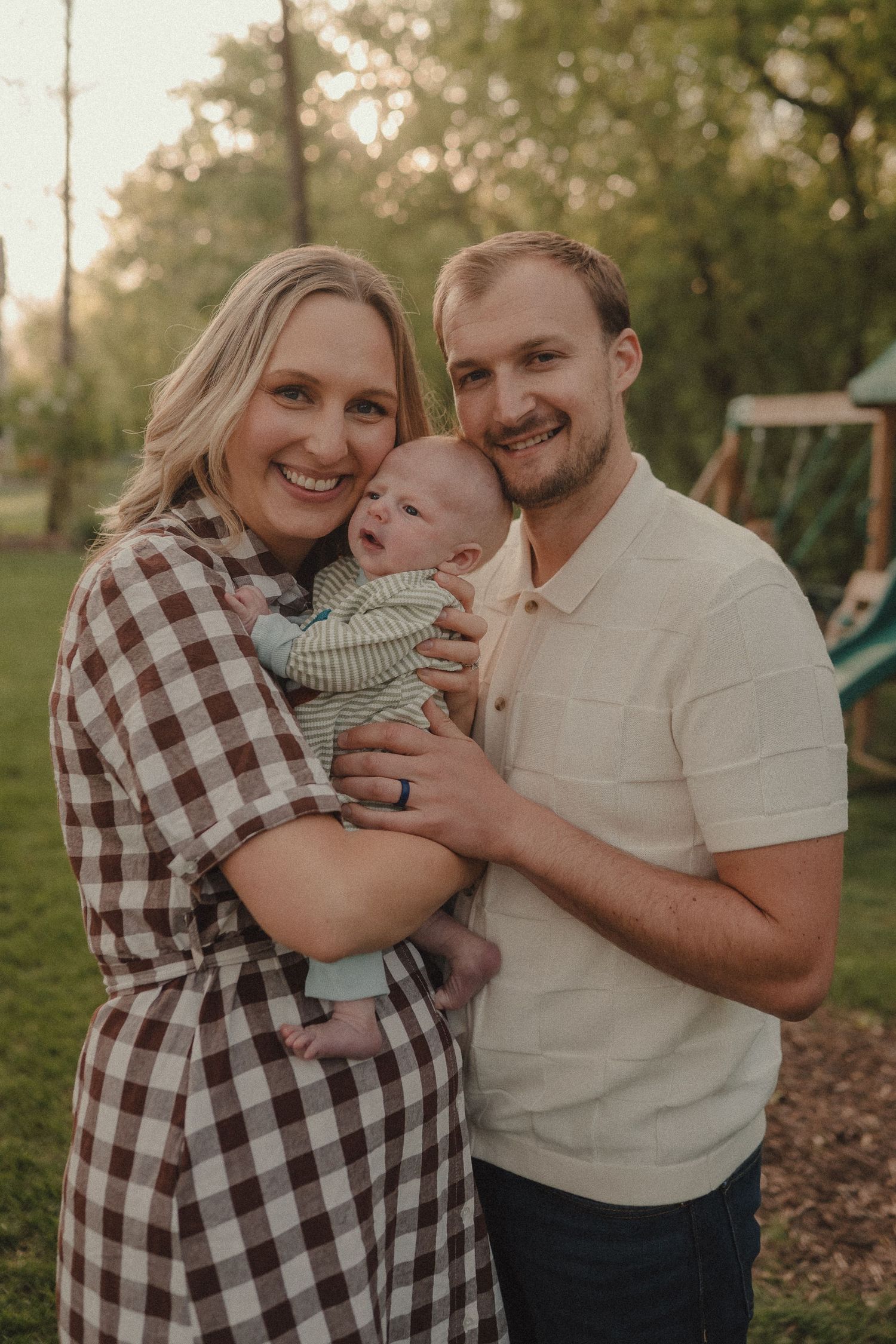 Happy young family with newborn baby posing outdoors in a sunny park setting.