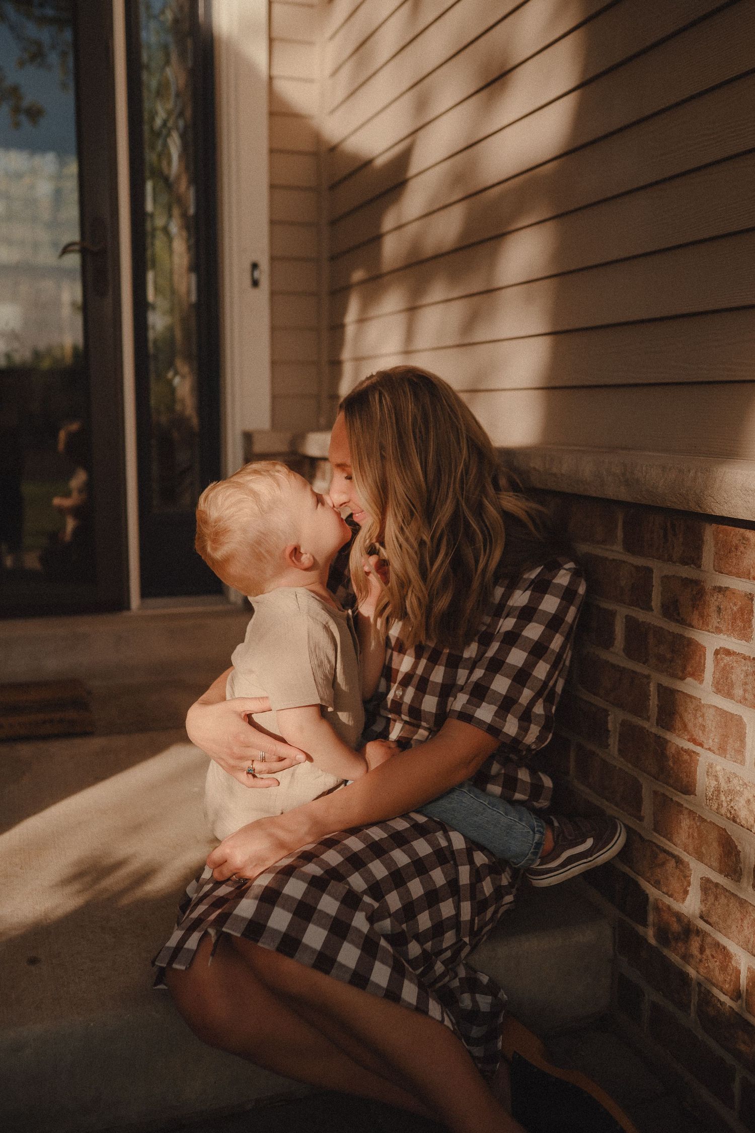 A loving mother in plaid dress sits on porch steps cuddling her toddler in warm golden sunlight.