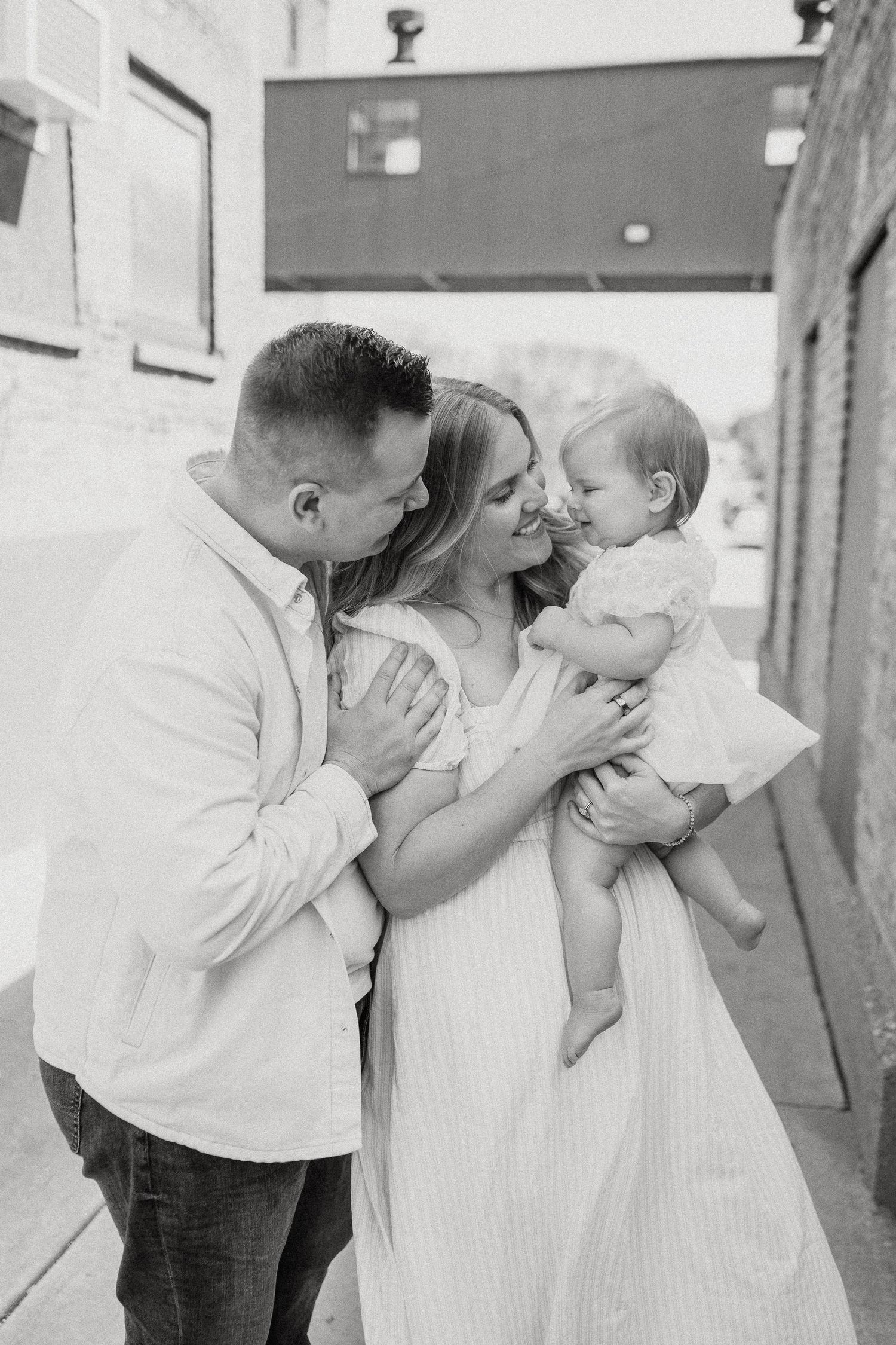 Black and white photo of a couple smiling at their baby girl in an urban alleyway setting.
