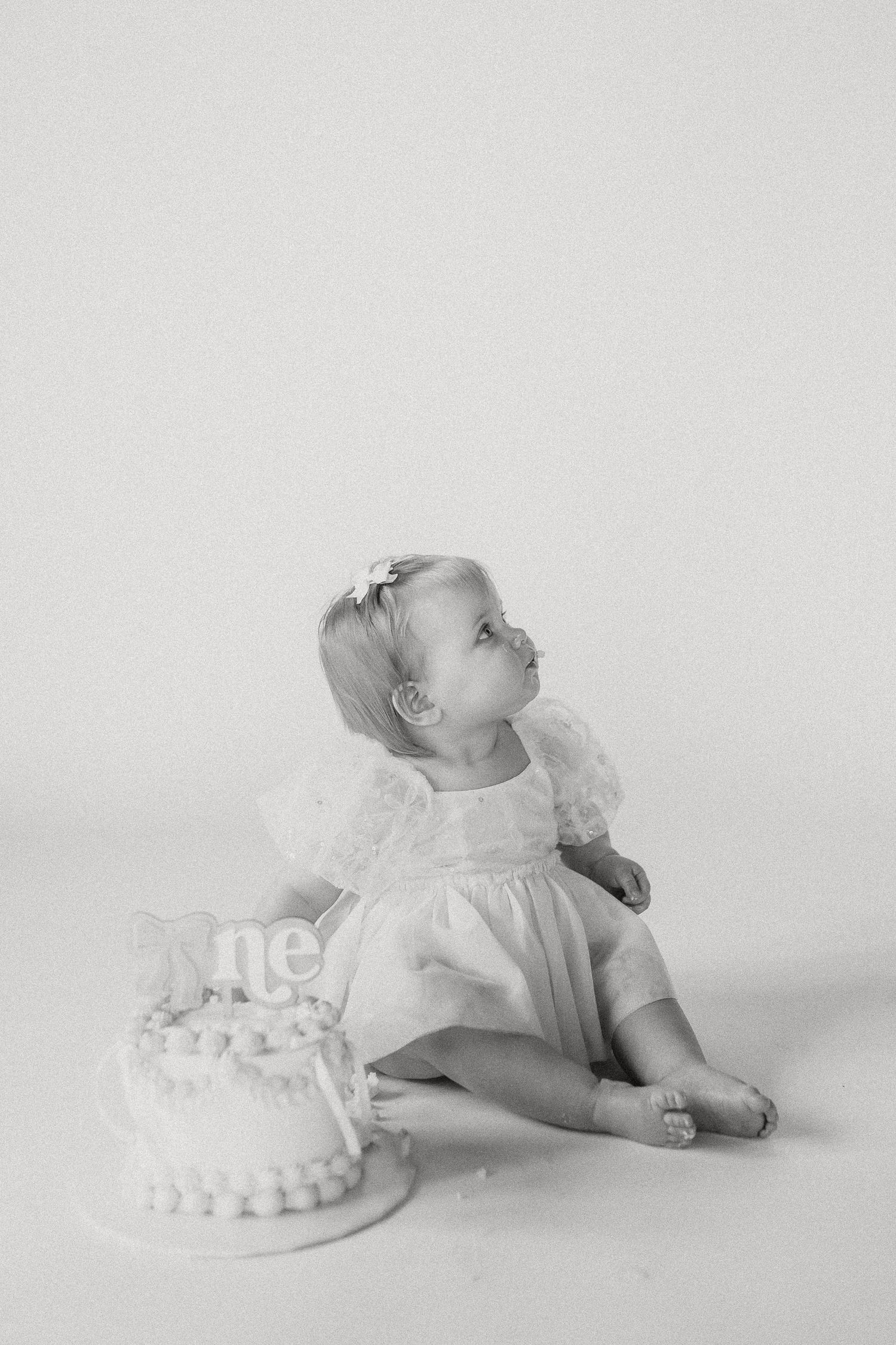 A baby girl in a white dress sits beside her first birthday cake in black and white photo.