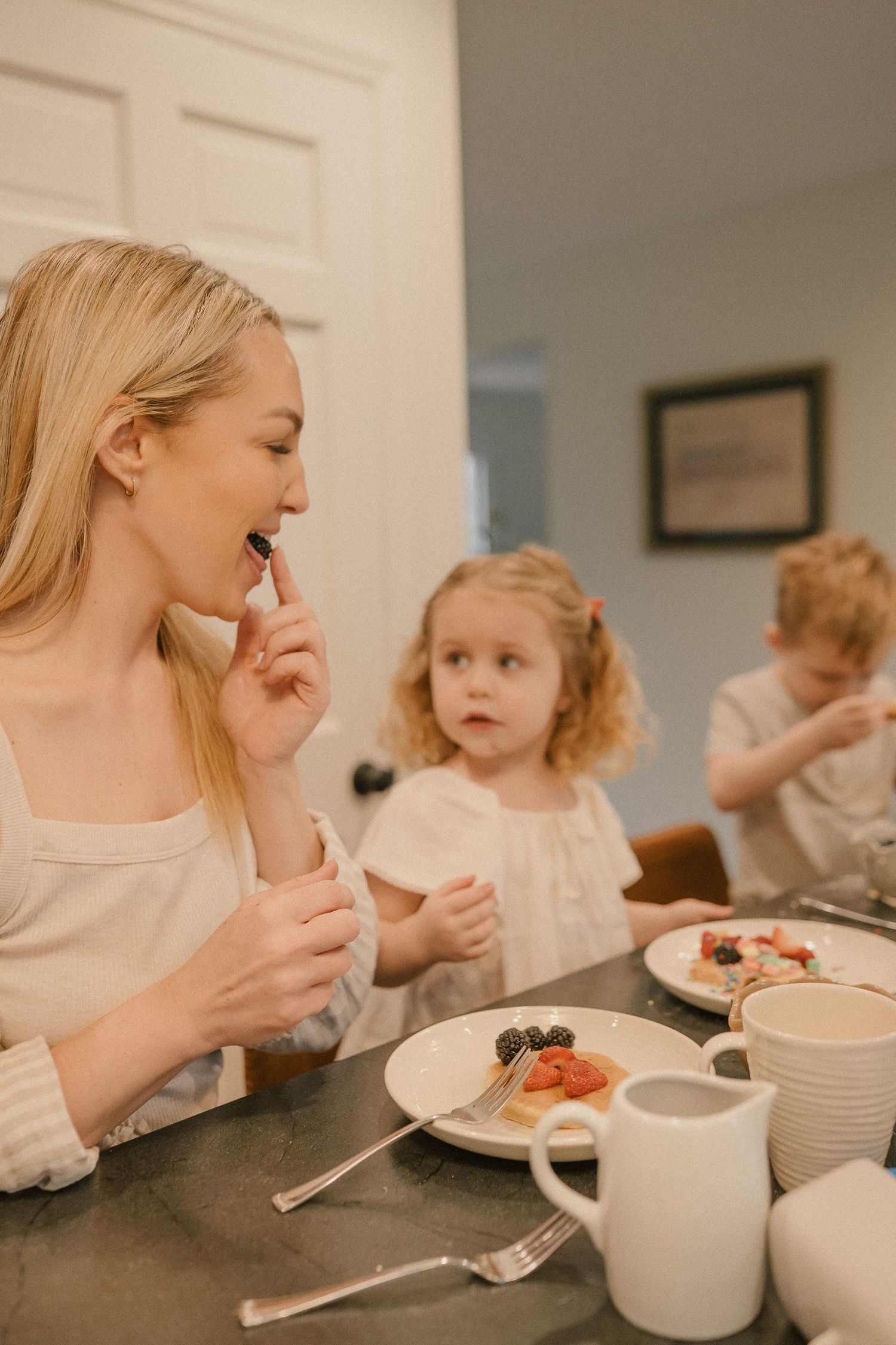 A smiling blonde mother eating a blueberry at a breakfast table with her two young children nearby.