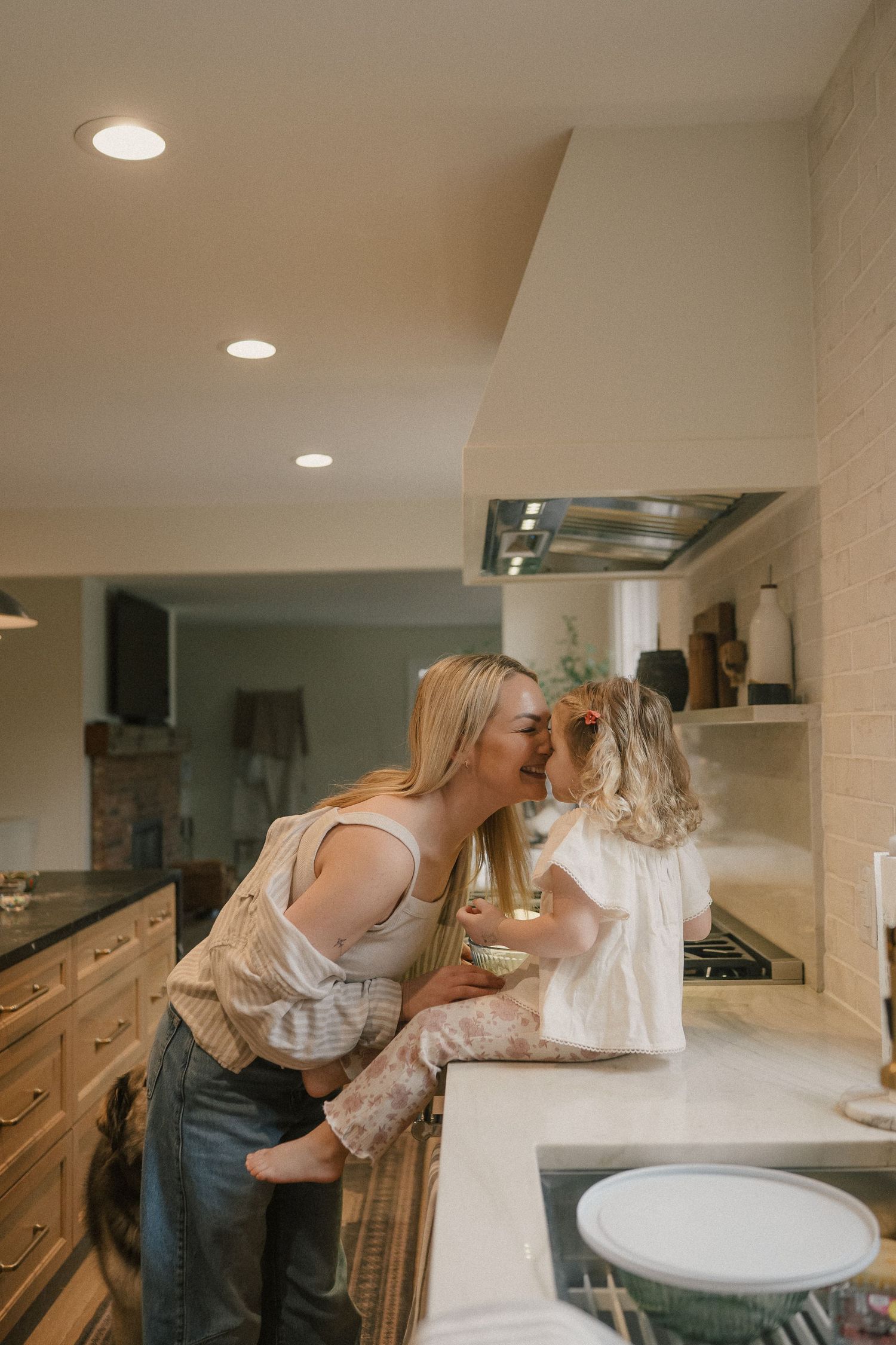Blonde mother kissing her young daughter while holding her in a bright modern kitchen.
