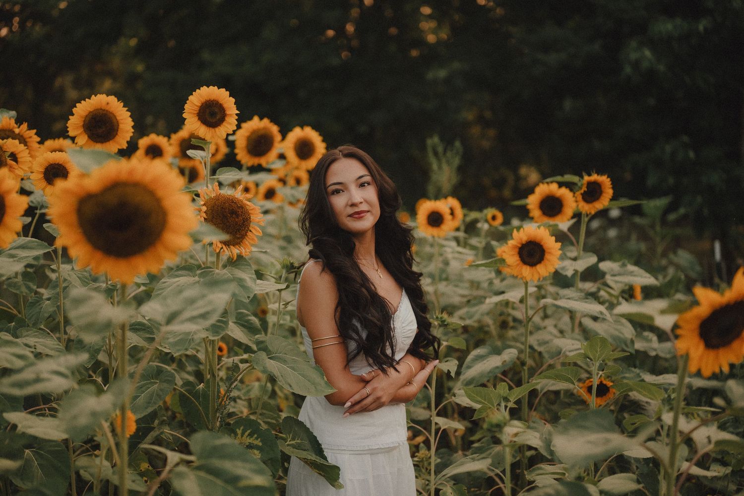 A young woman in a white dress poses gracefully among vibrant sunflowers in a lush garden at dusk.