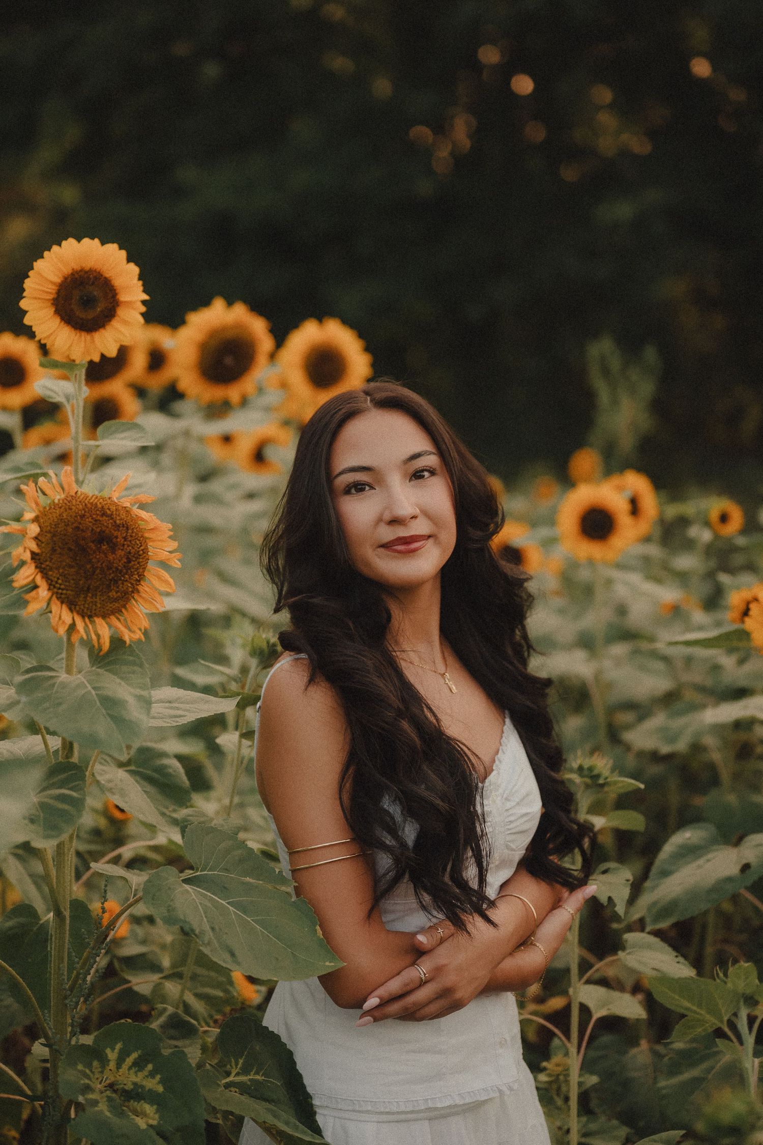 Young woman in white dress posing in sunflower field at sunset, arms crossed, long dark hair.