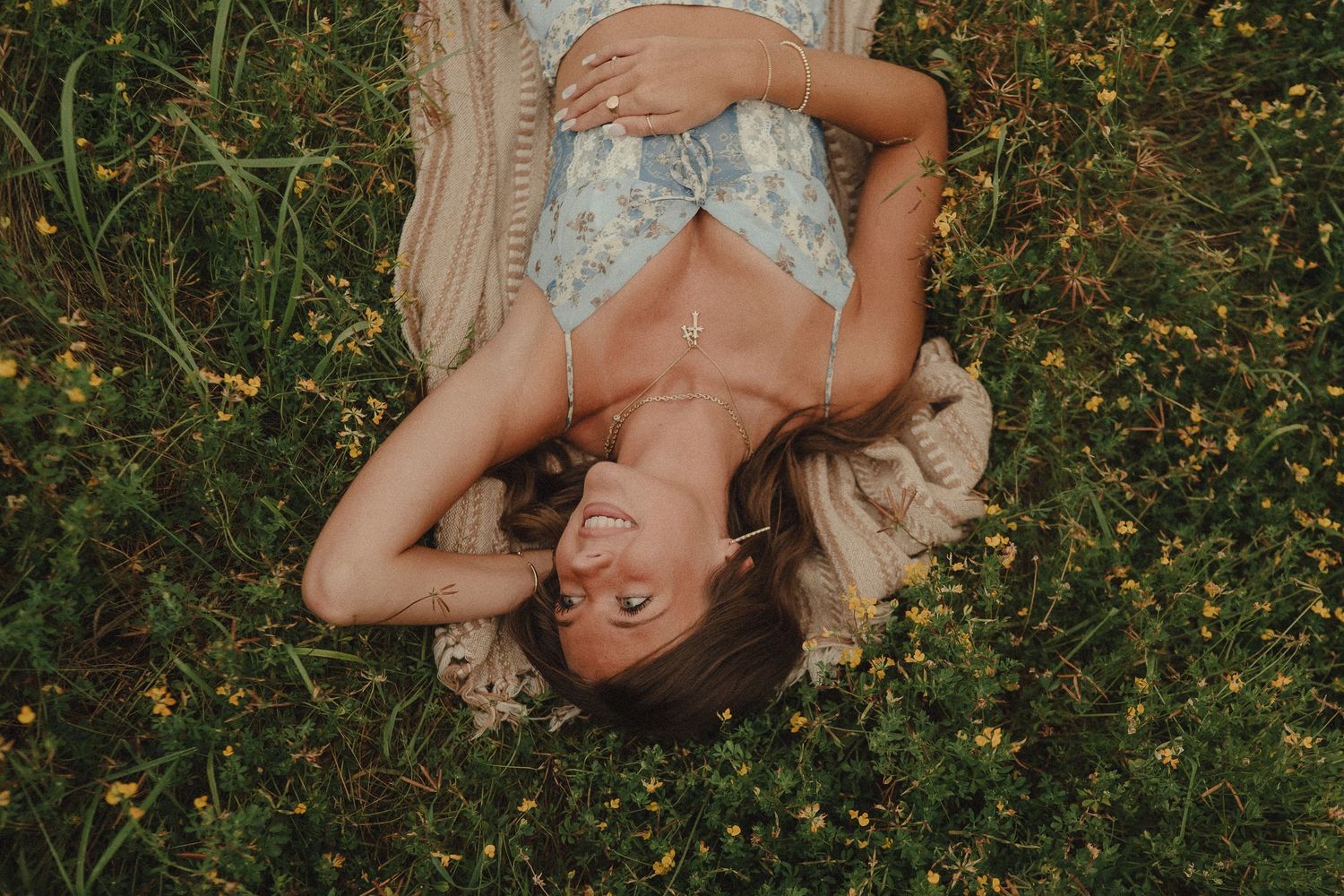 A joyful woman lying in a field of wildflowers, smiling while wearing a floral dress on a sunny day.