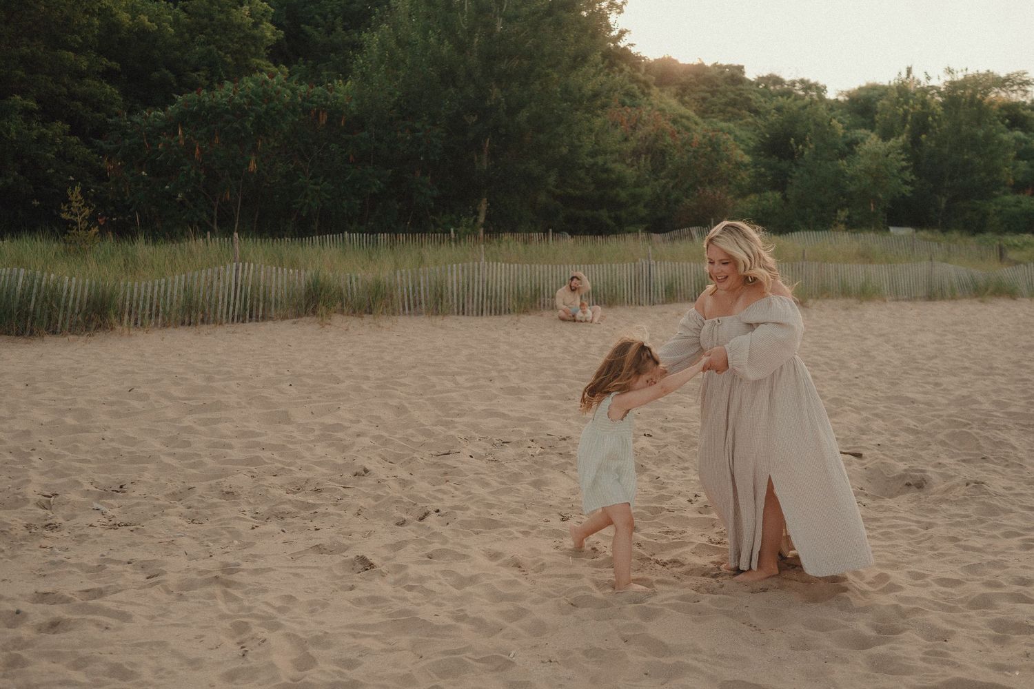 A mother in a flowing dress dances with her young daughter on a sunny sandy beach.