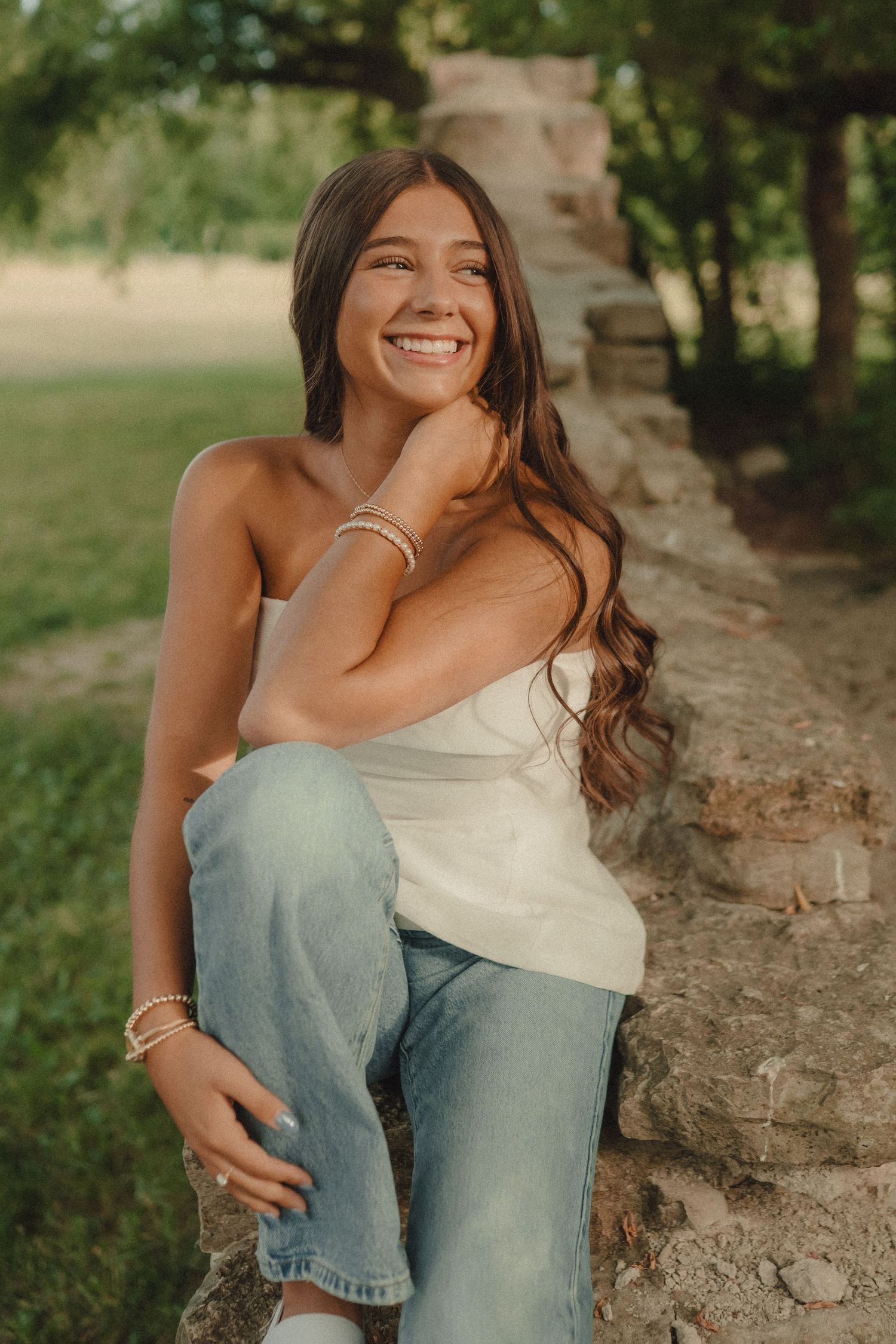 Smiling young woman in white tube top and jeans sitting on stone wall outdoors in green park setting.
