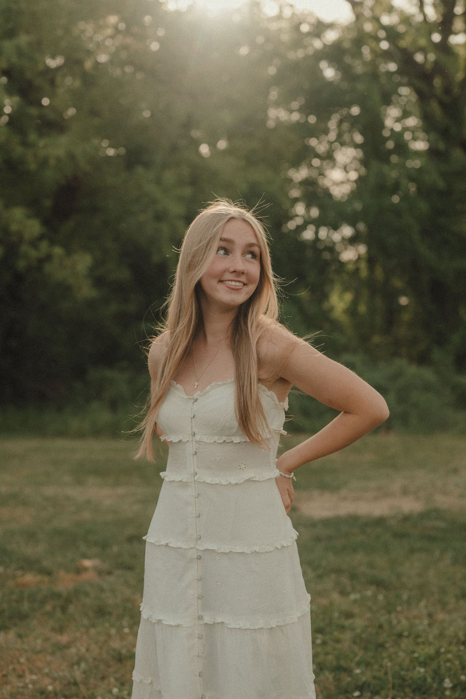 Young blonde woman in white sundress posing outdoors at golden hour with lush green trees in background.