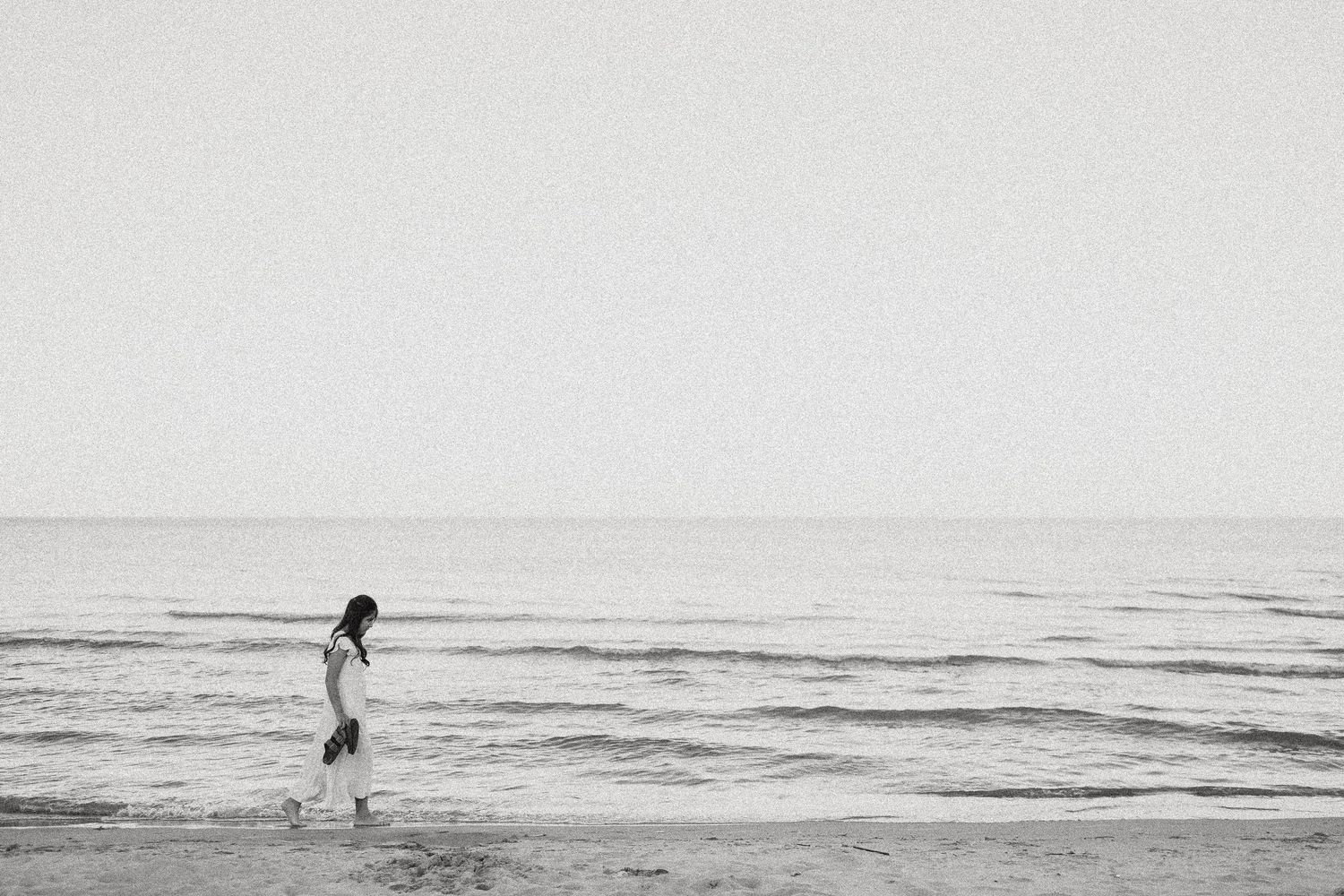 A lone woman walks along a serene beach shoreline in a black and white photo with calm ocean waves.