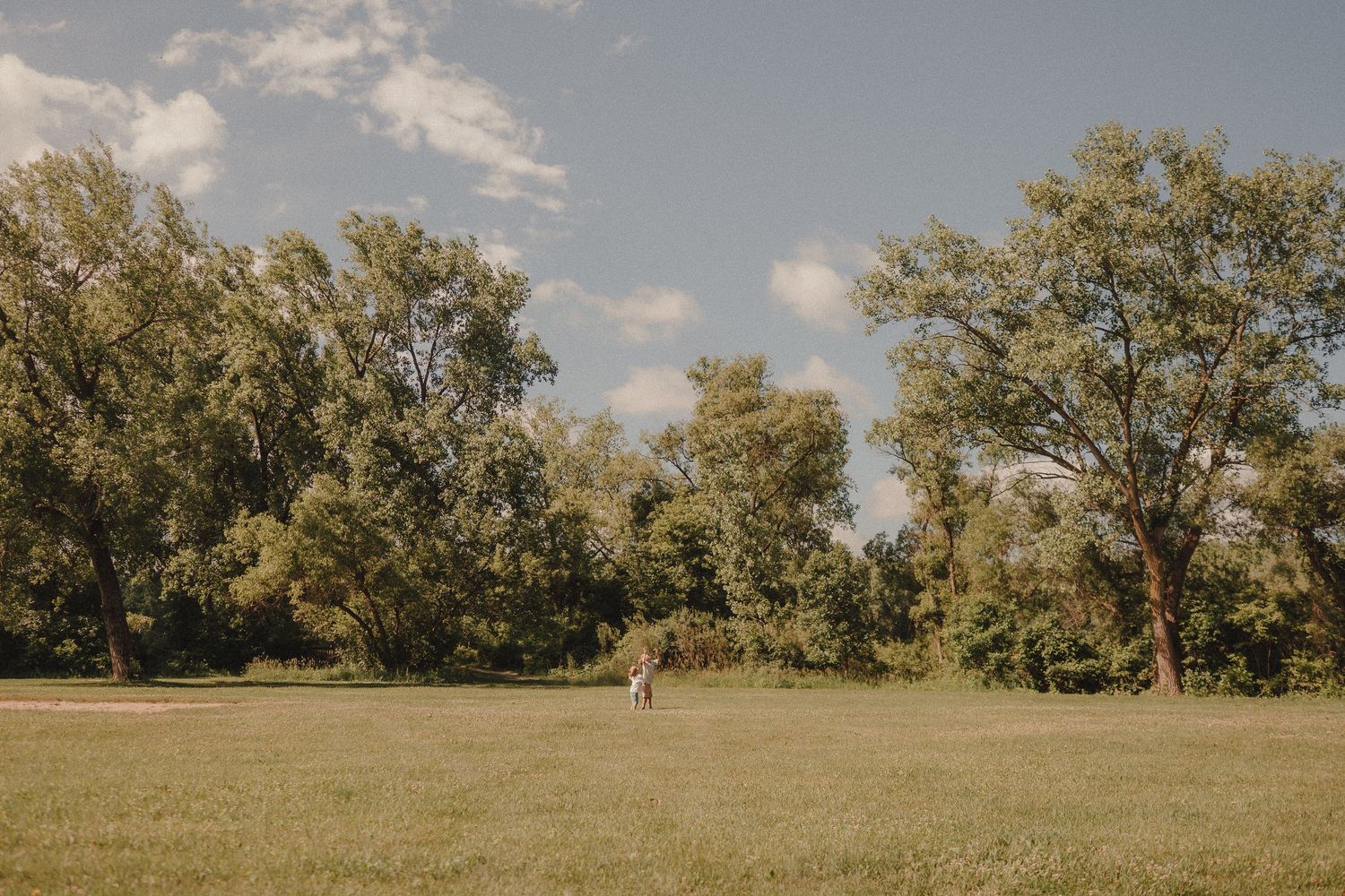 A couple stands in a lush green meadow surrounded by tall trees under a partly cloudy sky.