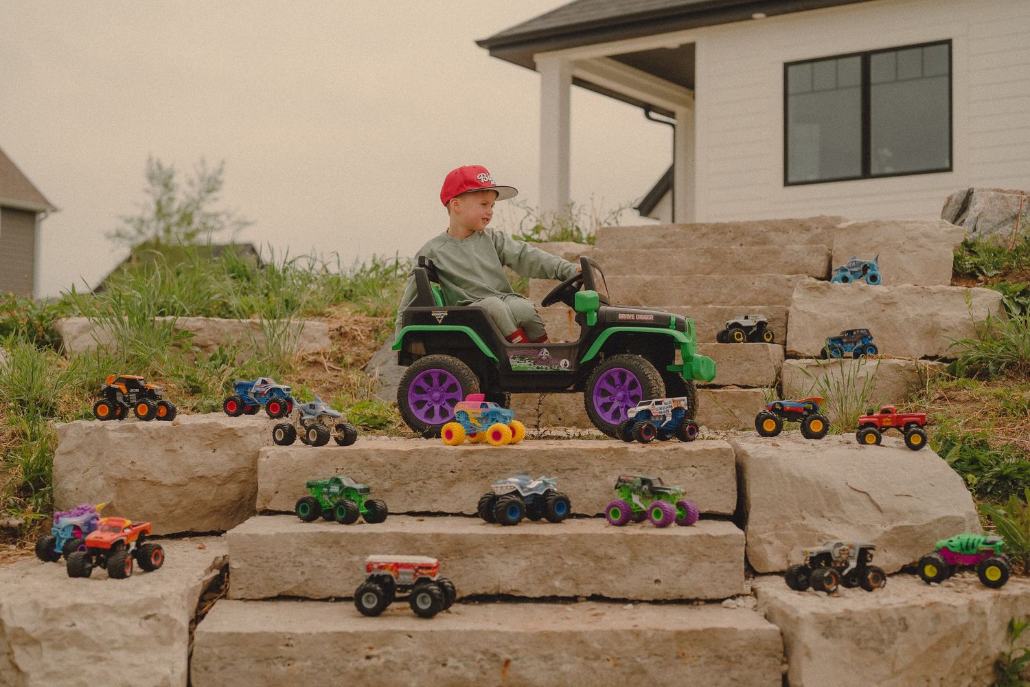Boy in red cap drives ride-on toy jeep surrounded by monster trucks on stone steps.
