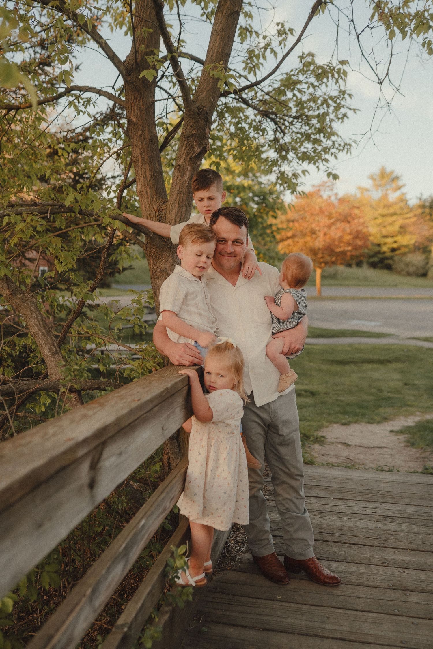 Father with four young children posing on wooden bridge surrounded by lush green trees at sunset.