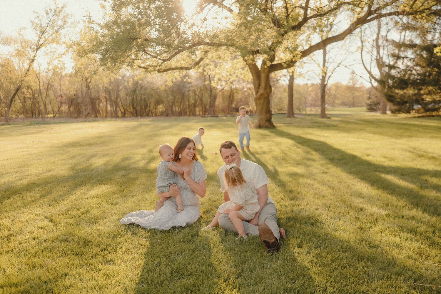 Family sitting on lush green grass under large oak trees during golden hour sunset in a park.