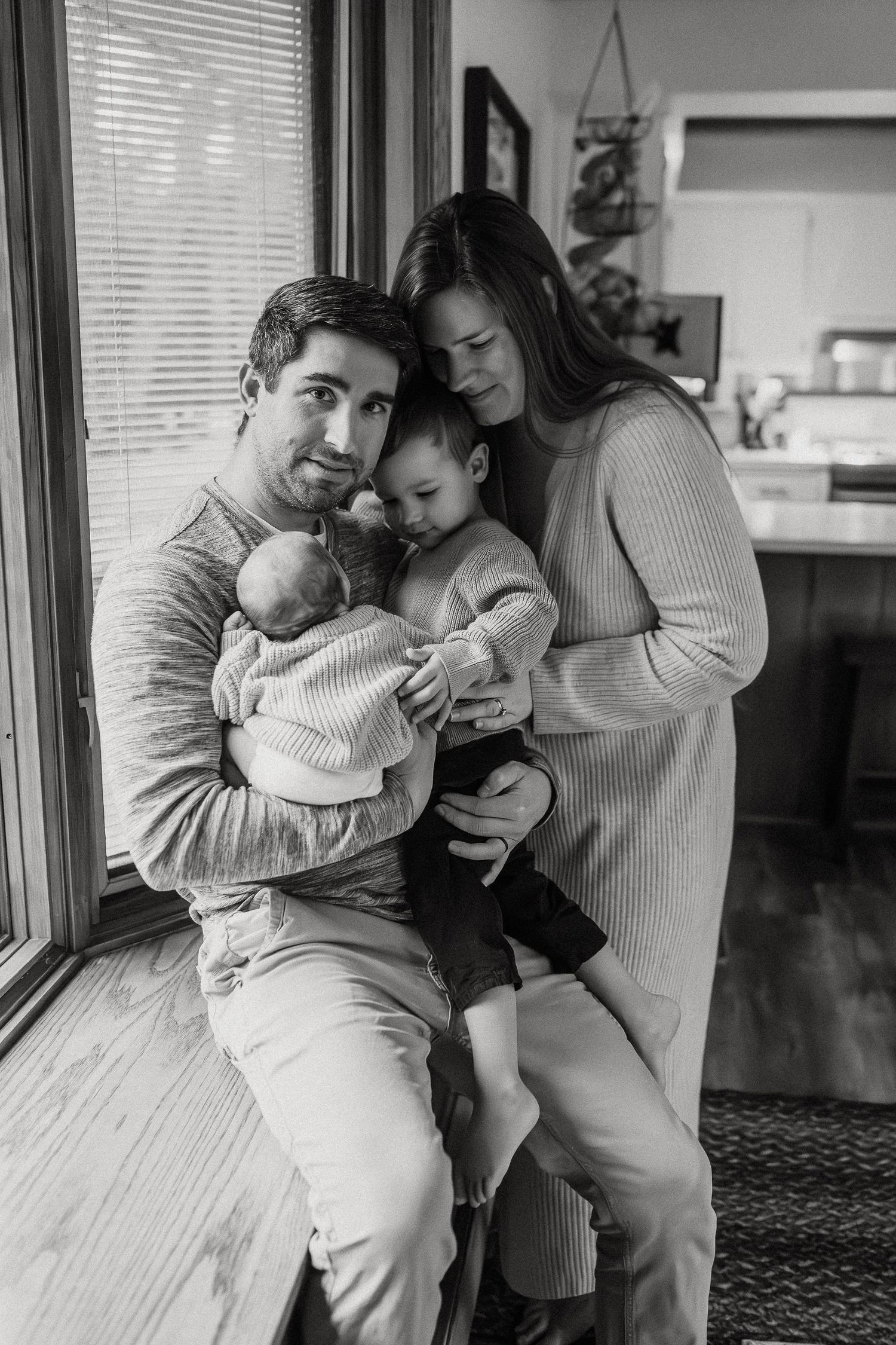A black and white photograph capturing tender family moments by a sunlit window.