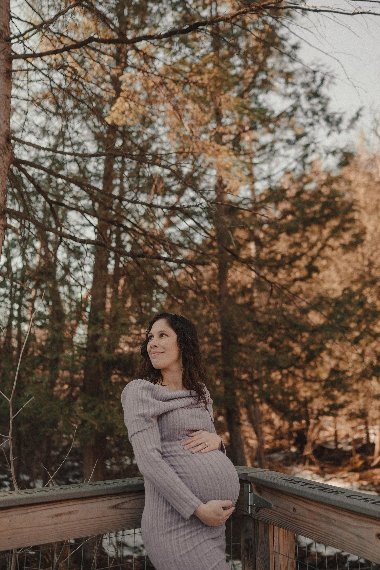 A pregnant woman in a knit sweater stands on a wooden deck surrounded by autumn trees at sunset.
