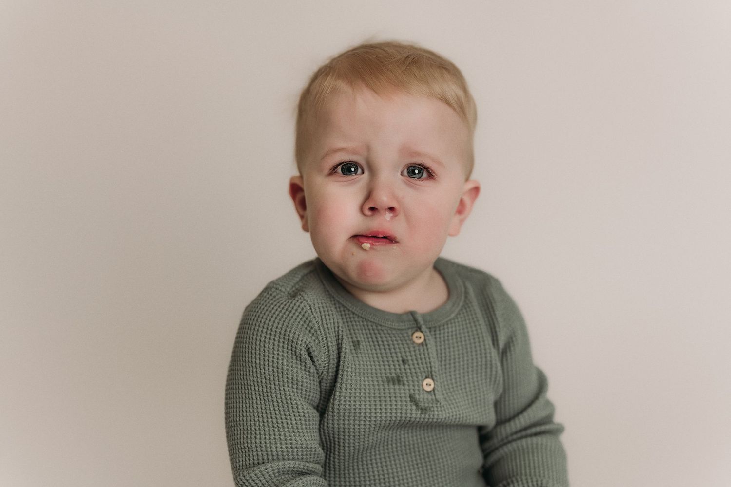 Baby in beige romper sits near wooden cake stand.