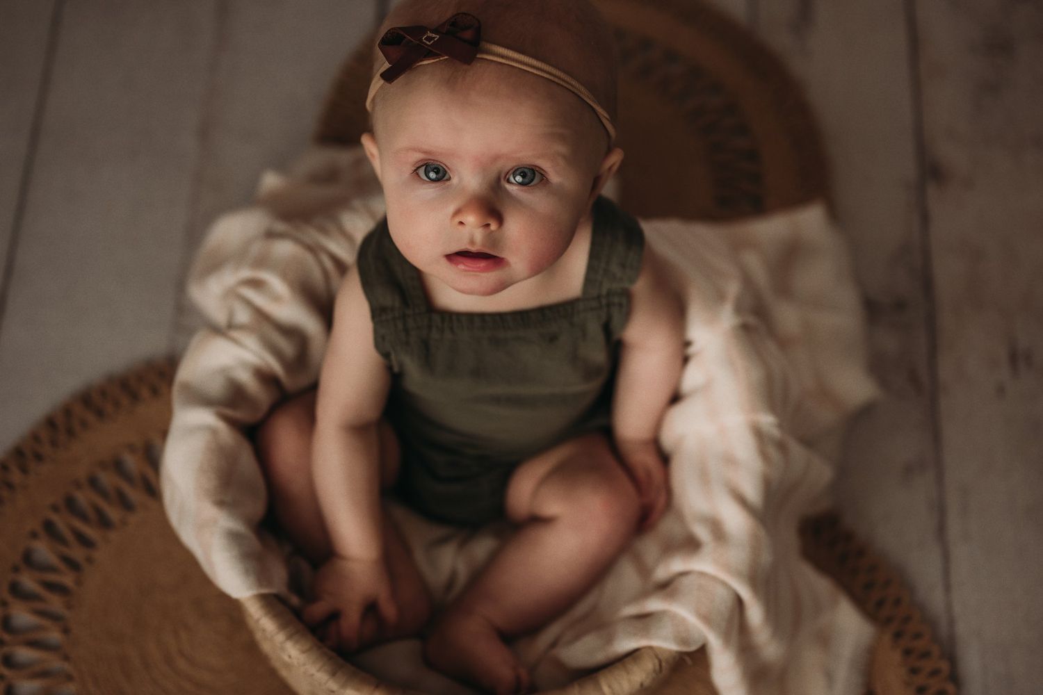 Baby in olive outfit sitting on circular woven mat.