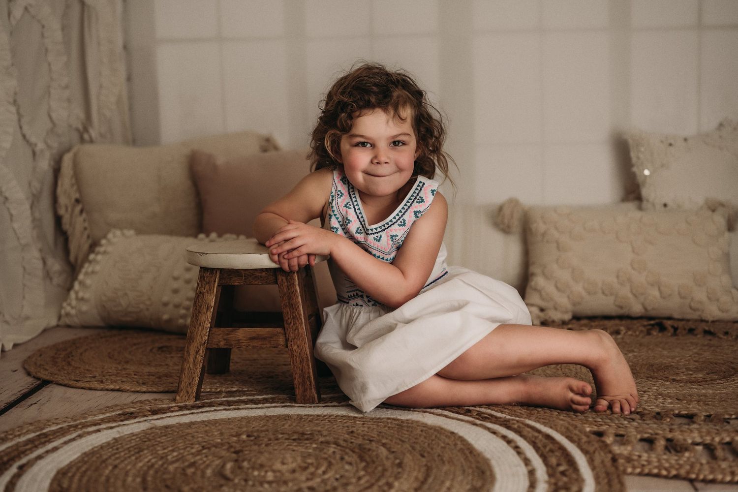 Child in casual dress leaning against wooden stool on circular patterned rug.