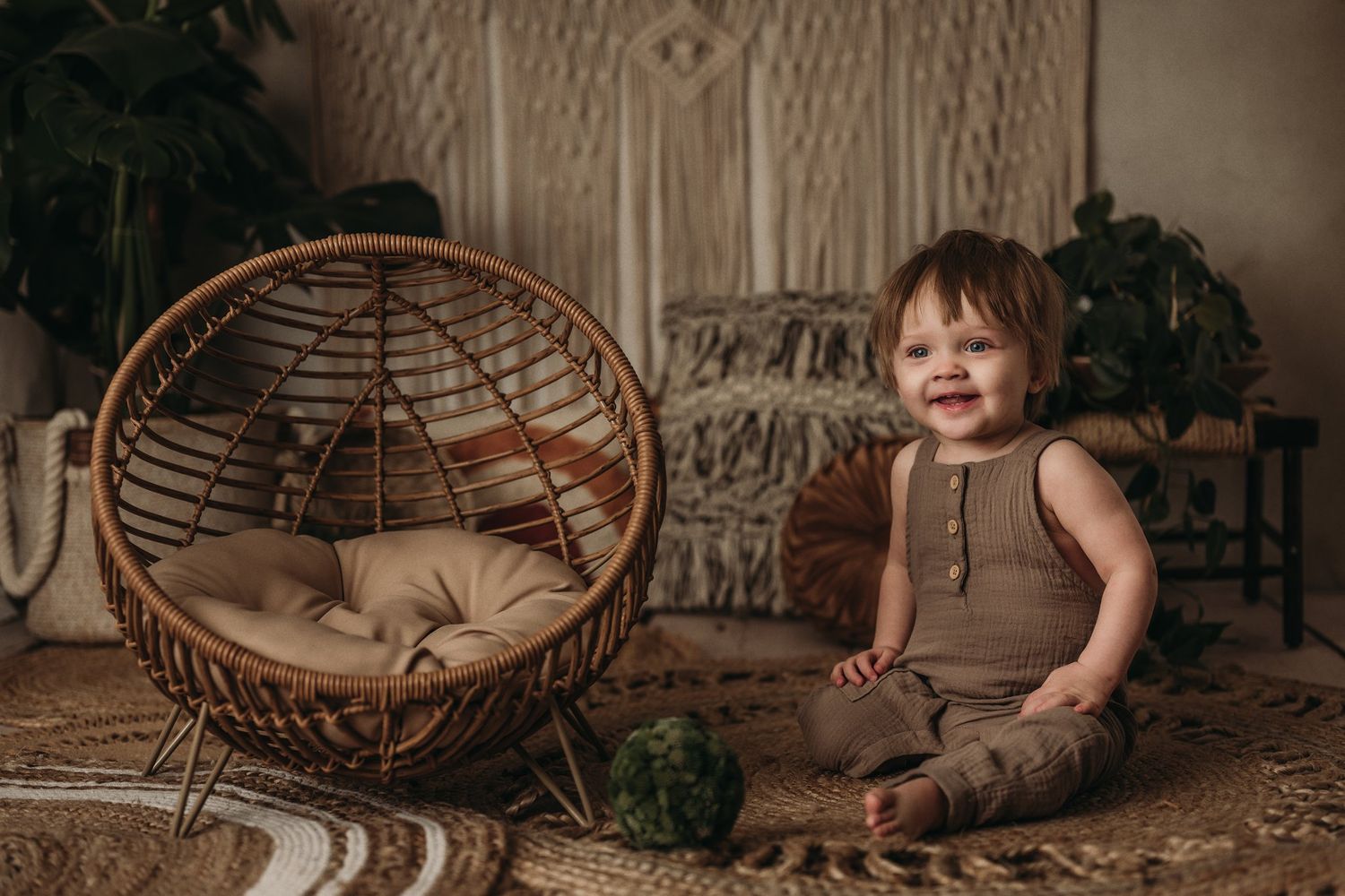 A small child sits beside a round rattan chair in a bohemian-styled room with macrame decorations.