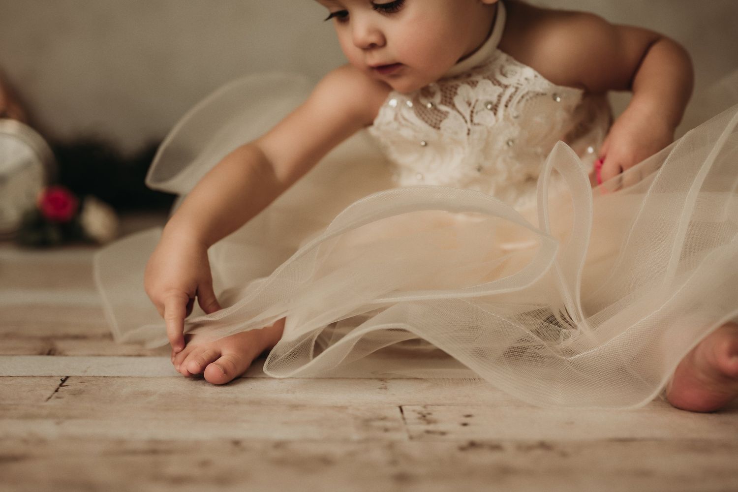 Tiny hands touch wooden flooring beneath flowing layers of delicate cream tulle fabric.