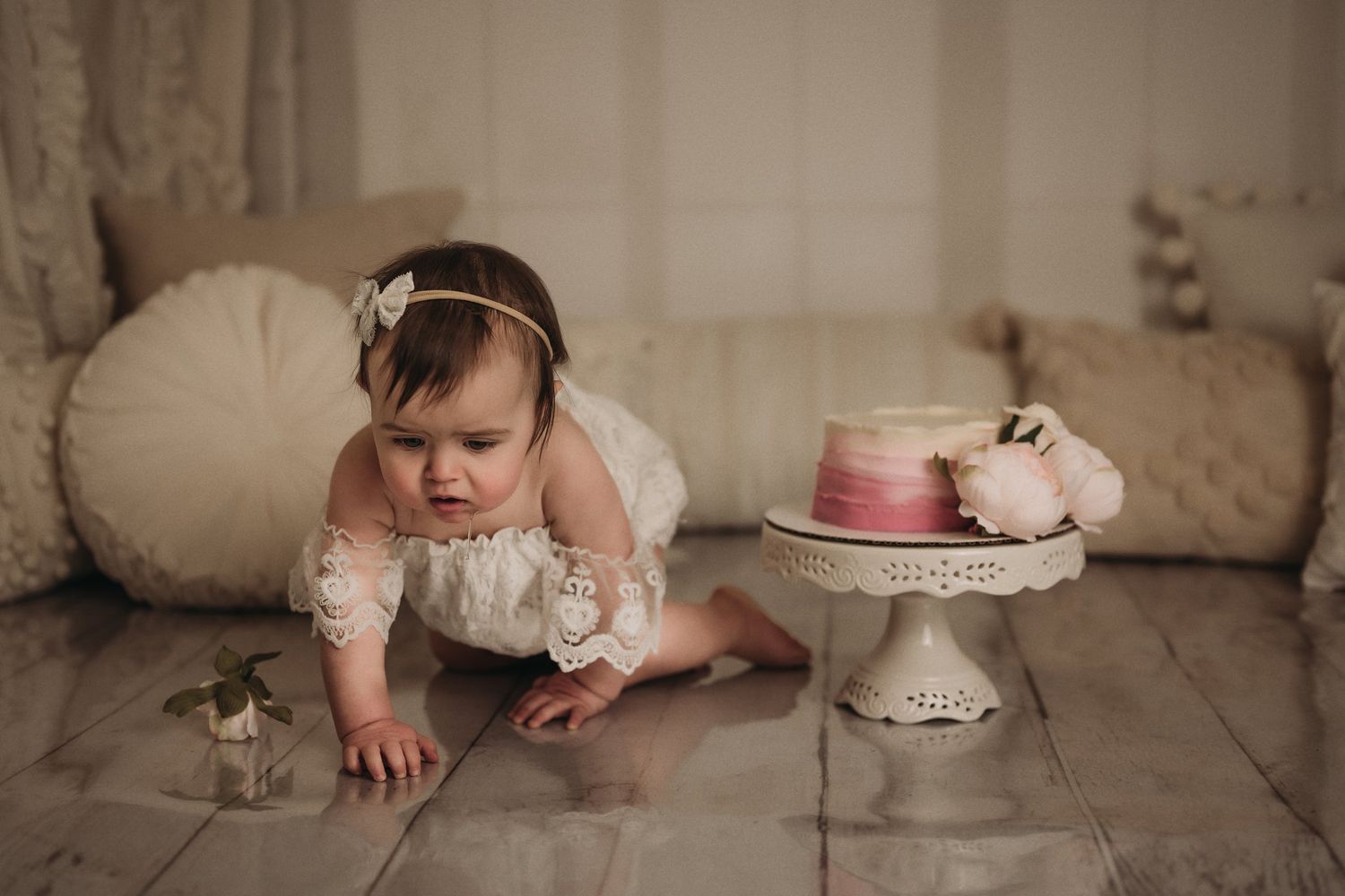 Baby in lace outfit crawling near pink cake on vintage cake stand with decorative pillows.