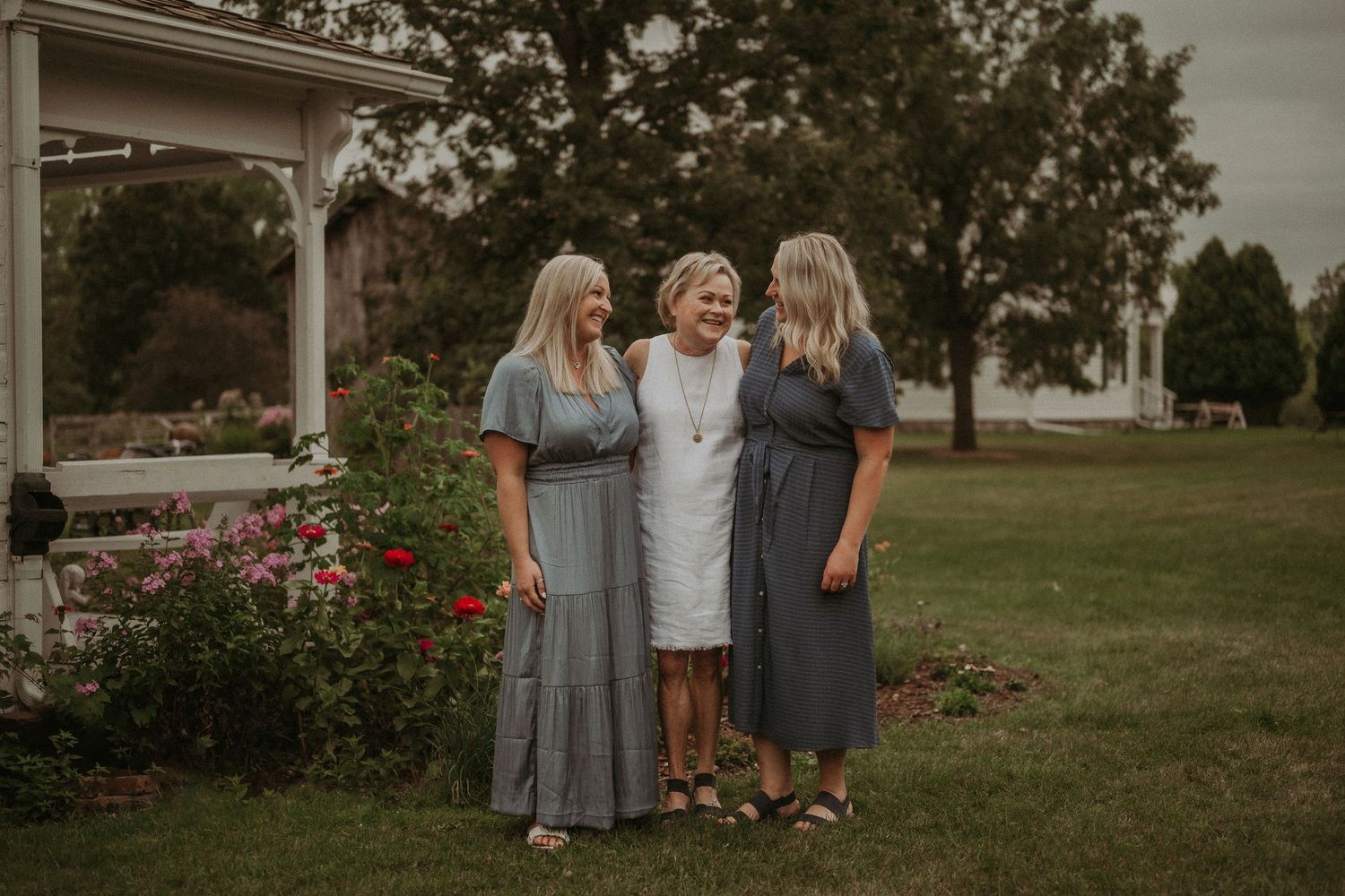 Three women in coordinating dresses stand together in garden with blooming red flowers.
