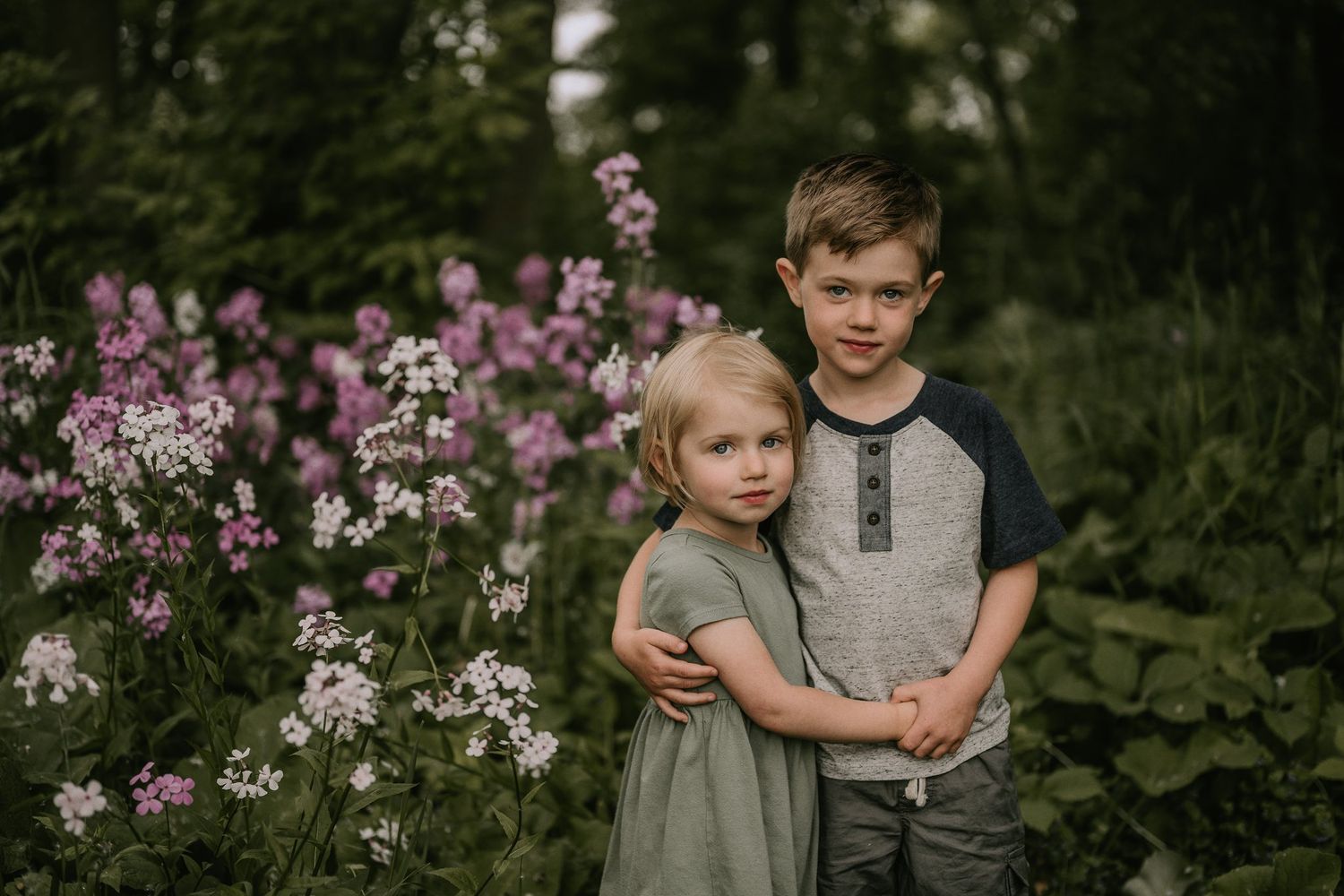 Two children stand together among blooming pink and white flowers in a natural outdoor setting.