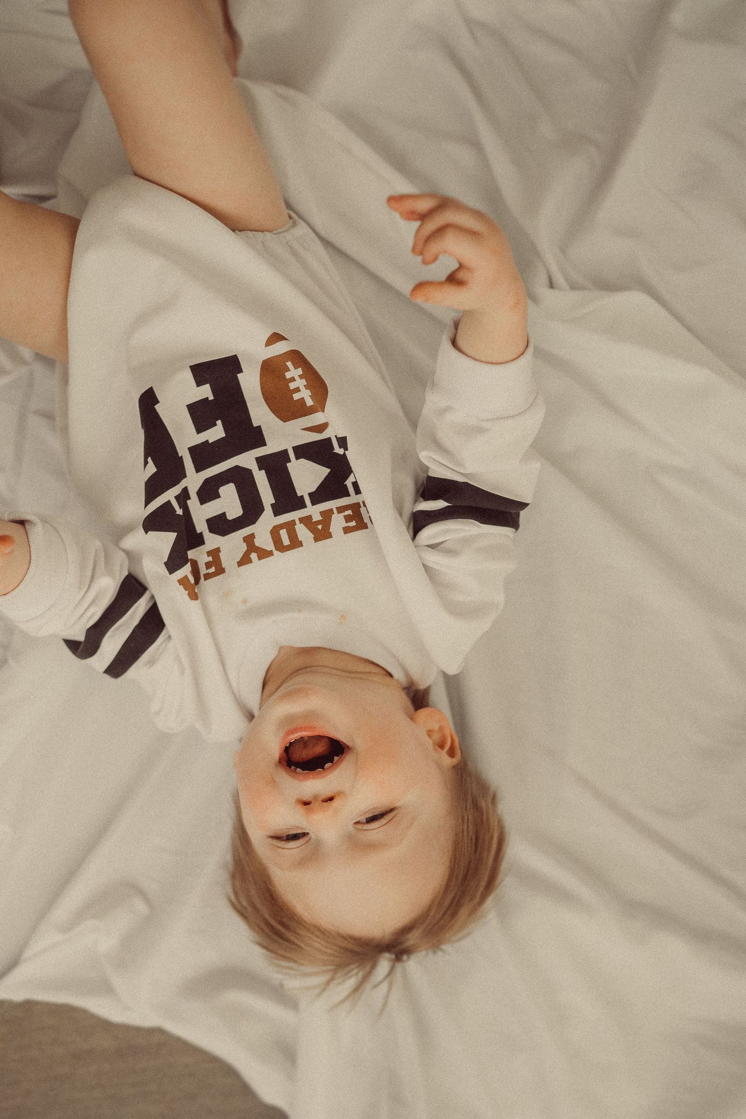 Playful overhead shot of toddler lying on white bedding wearing football themed bodysuit.