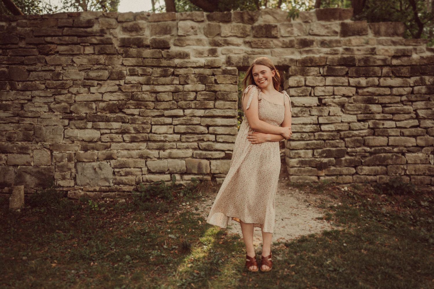 Woman in vintage-style dress poses elegantly against weathered stone wall in warm afternoon light.