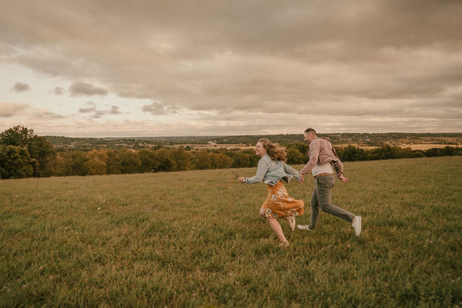 A joyful couple runs through a grassy hilltop field at sunset with dramatic cloudy skies overhead.