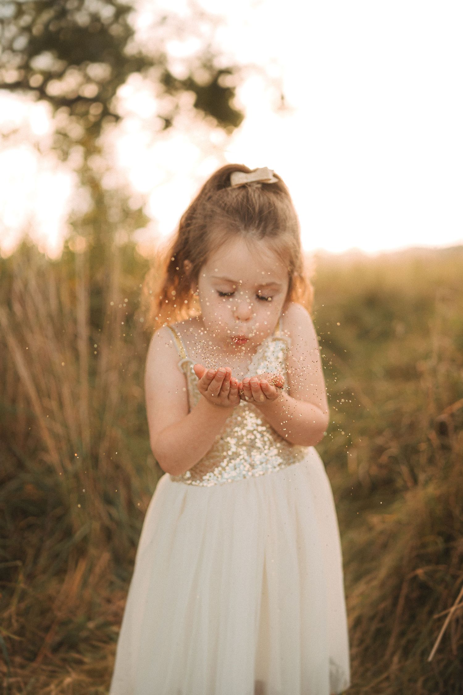 A child in a white dress blows glitter from their hands during golden hour.