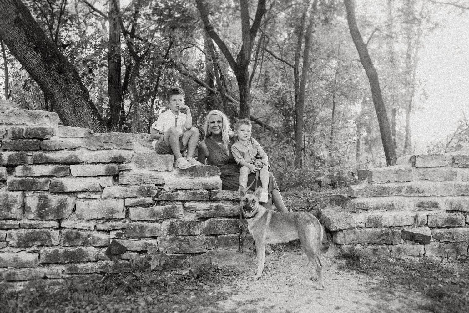 Black and white photo of a family and their dog sitting on old stone steps in a wooded area.