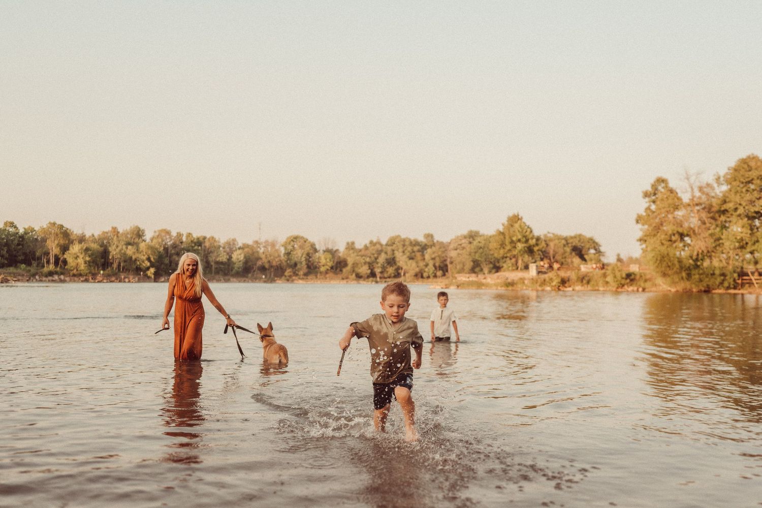 Family splashing and playing in a lake during golden hour with trees along the shoreline.
