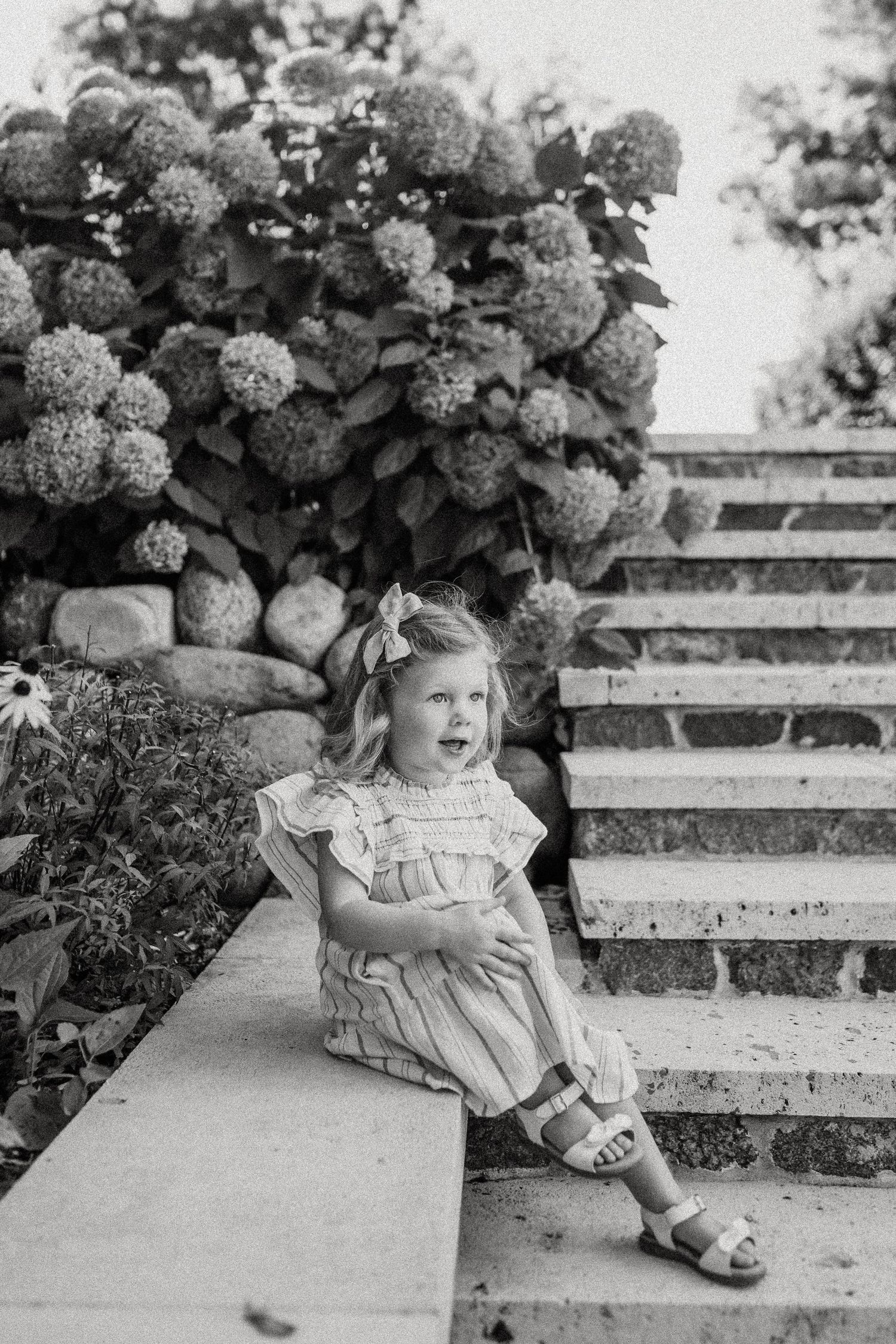 Black and white portrait of a child on stone steps surrounded by hydrangeas and rock walls.