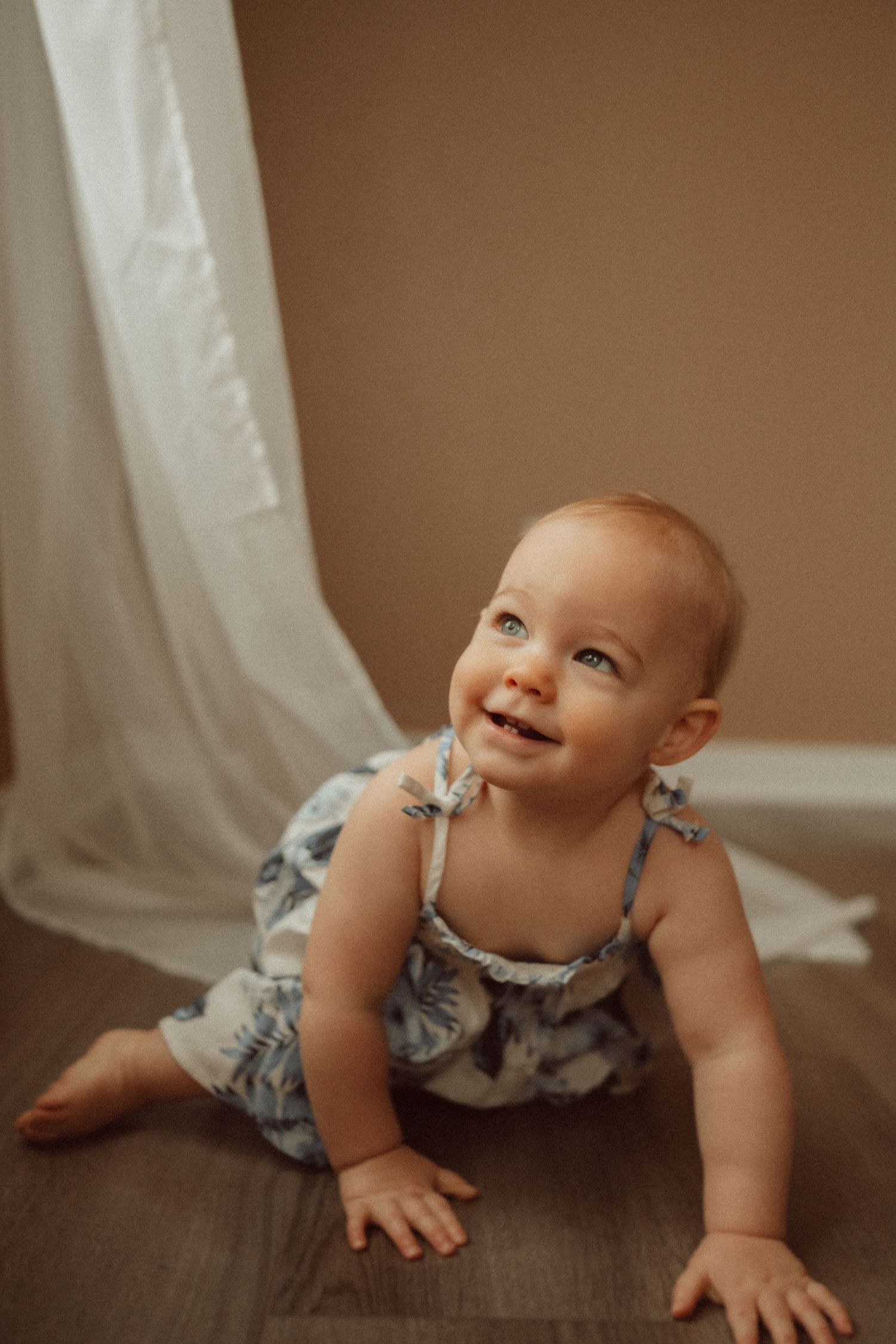 Baby in blue floral dress crawling on hardwood floor next to white curtain.