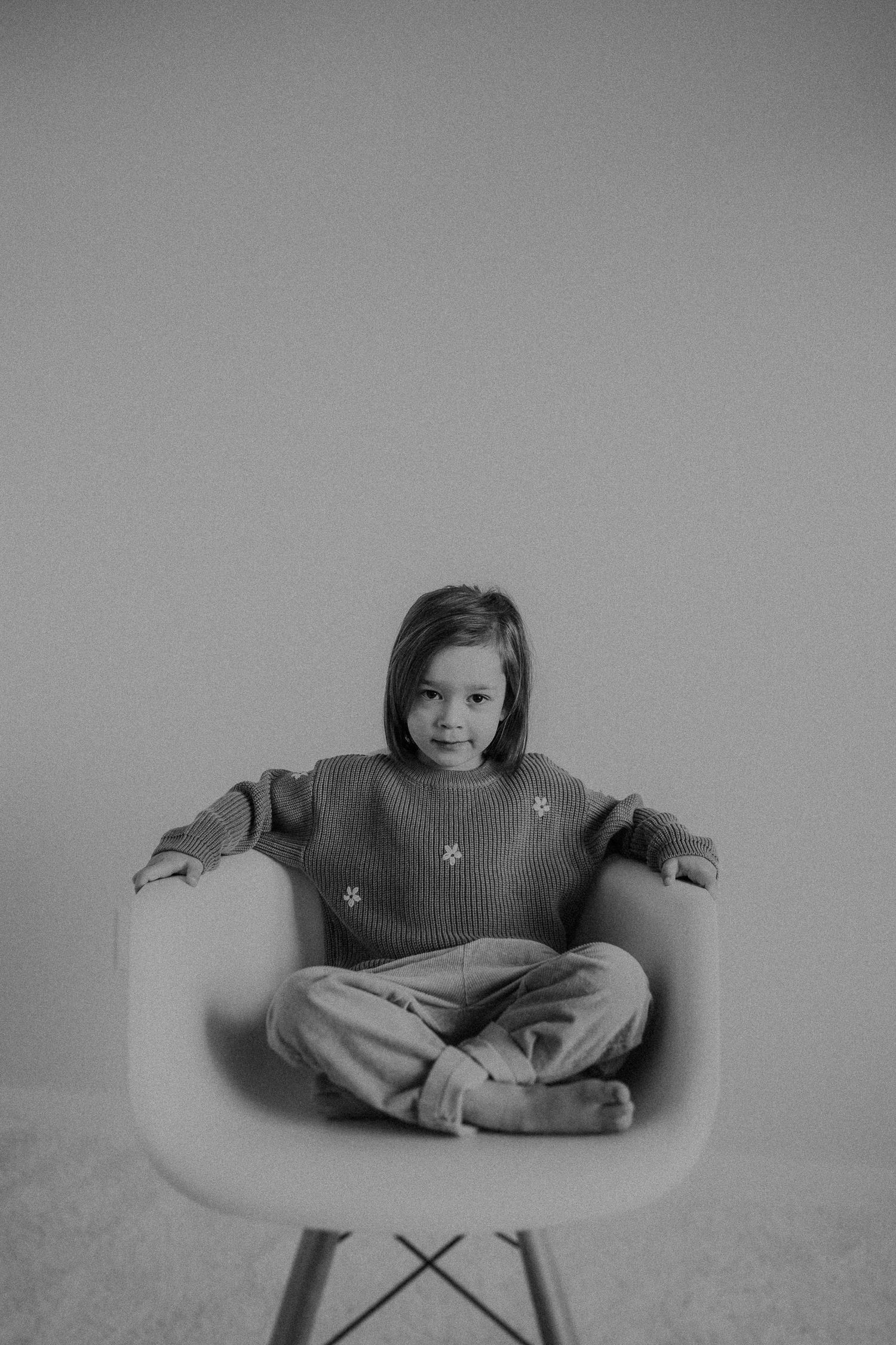 Child in knit sweater sitting cross-legged on white chair against neutral background.